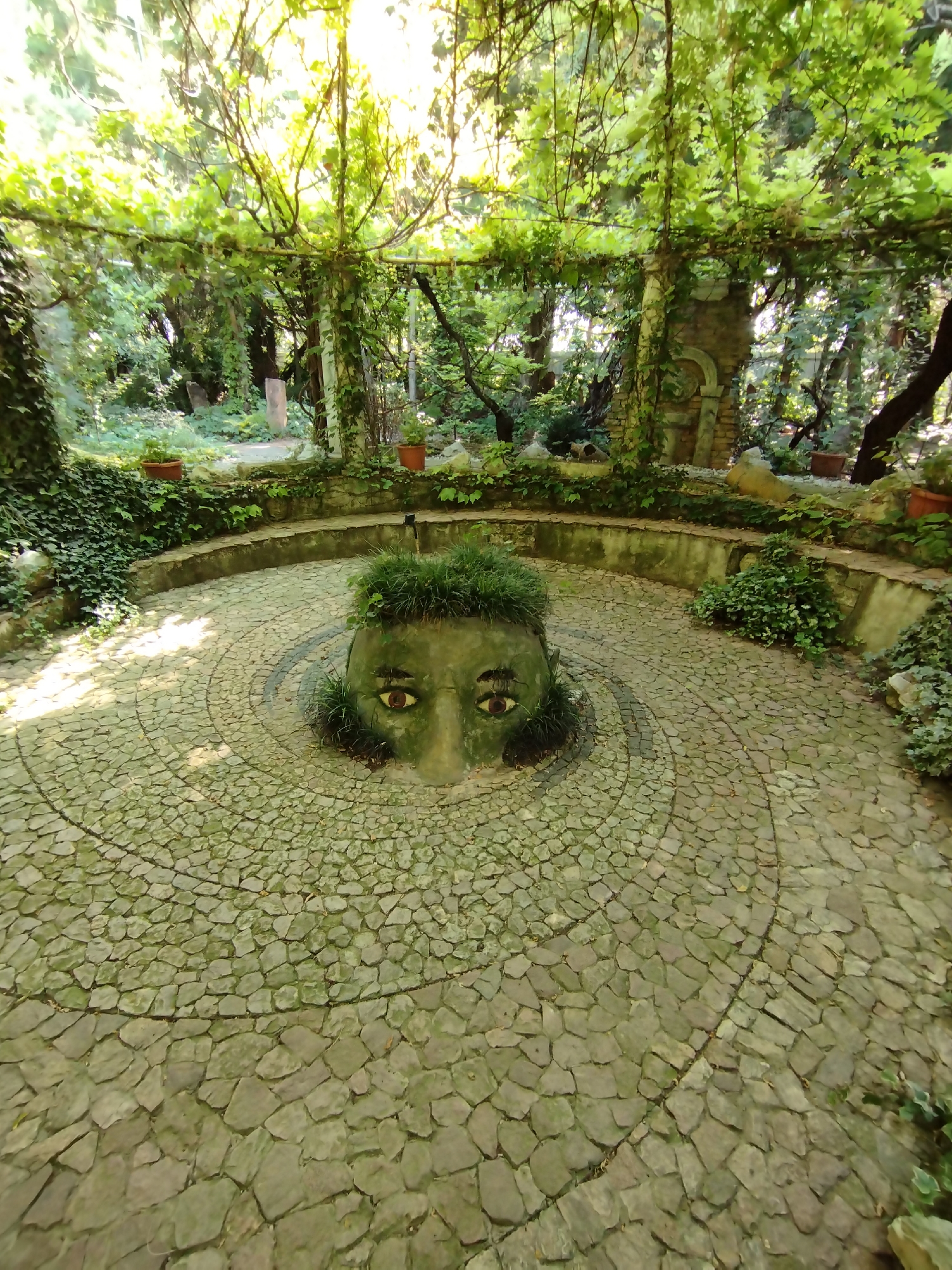 The image shows an outdoor stone-paved area with a sculpted head emerging from the ground in the center. The head appears to be made of stone or concrete and is partially covered in grass, resembling hair. It has large, prominent eyes with distinct pupils. The surrounding area is a circular patio paved with irregular, light-colored stones. Behind the patio, there is a curved, low stone wall overgrown with ivy and other greenery. The background consists of a lush, dense forest with trees and foliage, suggesting a natural or semi-natural setting. The lighting indicates it is daytime, with sunlight filtering through the trees, casting dappled shadows on the ground. The overall scene evokes a sense of a hidden or artistic garden space. The location in Kattasu, Uzbekistan, is not visually evident from the image itself.