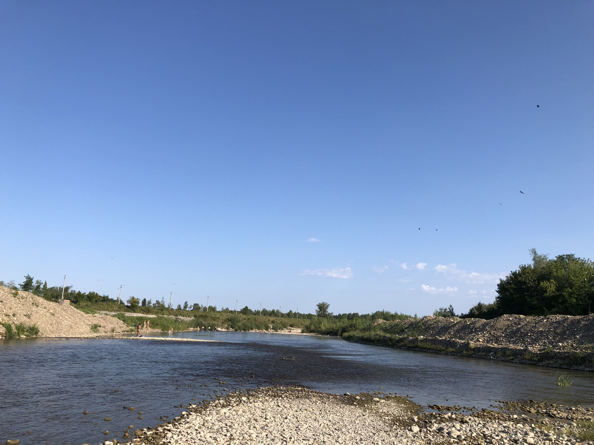 This is a wide shot of a river with a clear blue sky above. The river is shallow and rocky, with a small sandbar in the foreground. On the far bank, there is lush green vegetation and trees. Several people are visible in the water and on the riverbanks. The scene appears to be a natural environment, likely a recreational area within the city of Nyzhnii Verbizh, Ukraine.