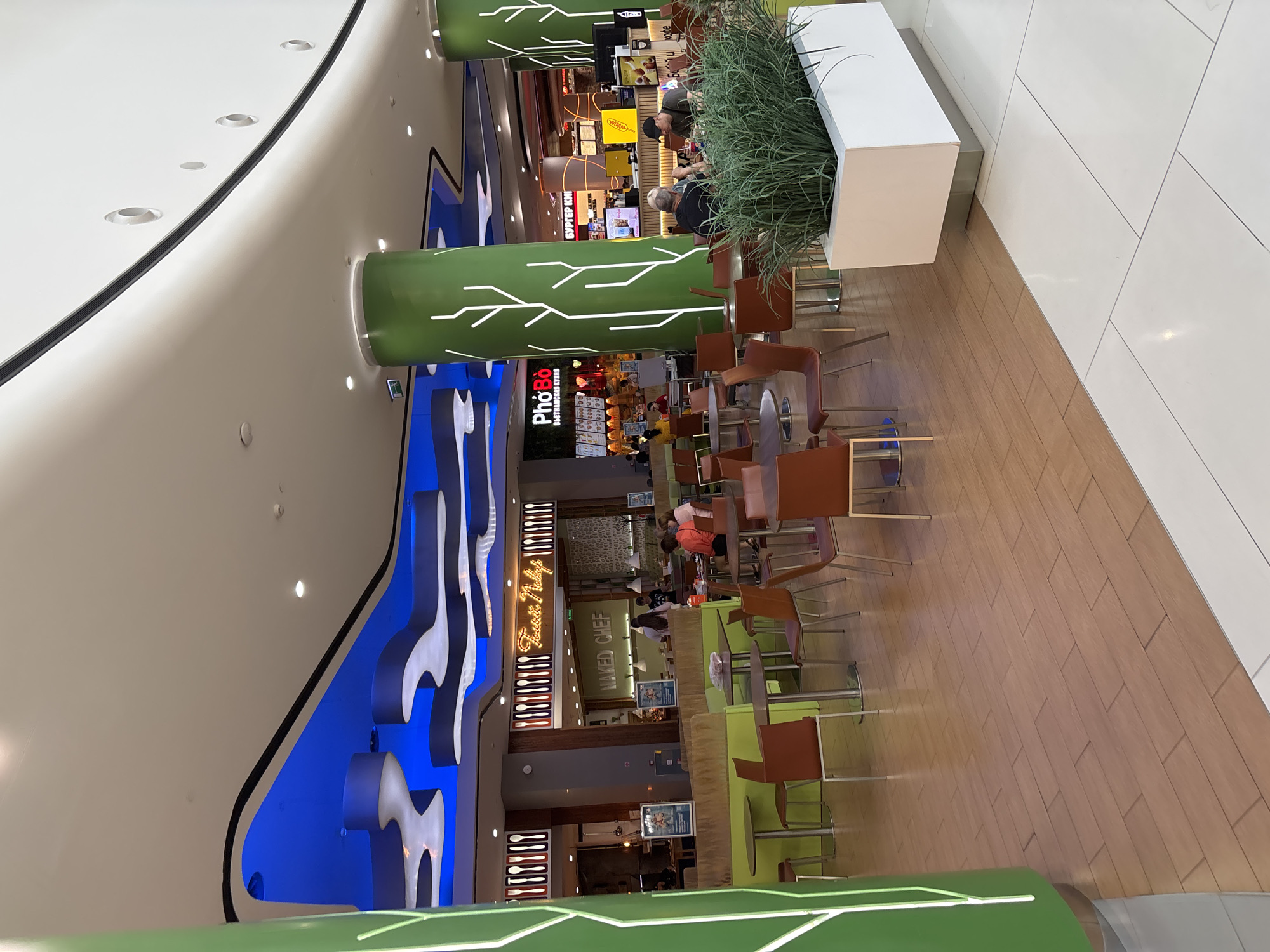 The image depicts a food court area within a shopping mall, featuring a vibrant and modern design. Large, green, cylindrical pillars with white tree-like graphics are a prominent element of the interior architecture. The ceiling is adorned with a blue, wave-like design, creating a dynamic visual effect.
Several food stalls are visible, including "PhoBo" and "Naked Chef," suggesting a variety of culinary options. Tables and chairs, predominantly in brown and orange hues, are arranged throughout the space, with some occupied by patrons. The flooring is a mix of wood-look tiles and white tiles. A white planter box filled with lush green foliage is placed in the foreground, adding a touch of nature to the urban setting. The overall atmosphere appears to be casual and bustling, typical of a food court environment. The location of Tlustenkhabl, Russia, is noted as the setting, though no specific landmarks or identifiers within the image confirm this directly.