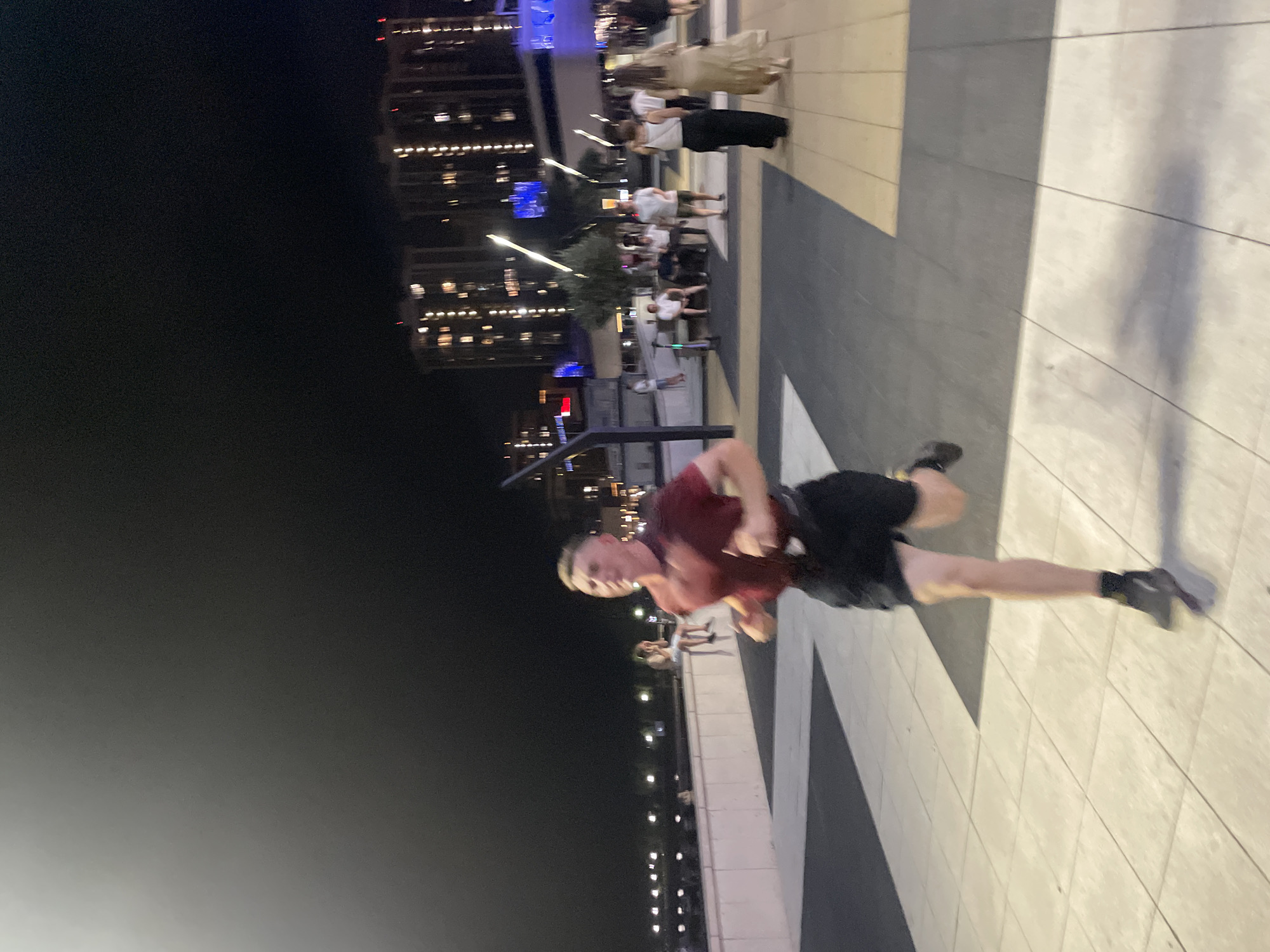 A young Caucasian male is captured mid-stride while running on a tiled pathway at night. He is wearing a dark red t-shirt and black shorts, with dark athletic shoes. The pathway is illuminated by artificial lights. In the background, several people are visible walking and sitting, and some multi-story buildings with illuminated windows line the horizon. The sky is dark, indicating it is nighttime. The location is the city of Sadovyi, Russia.
