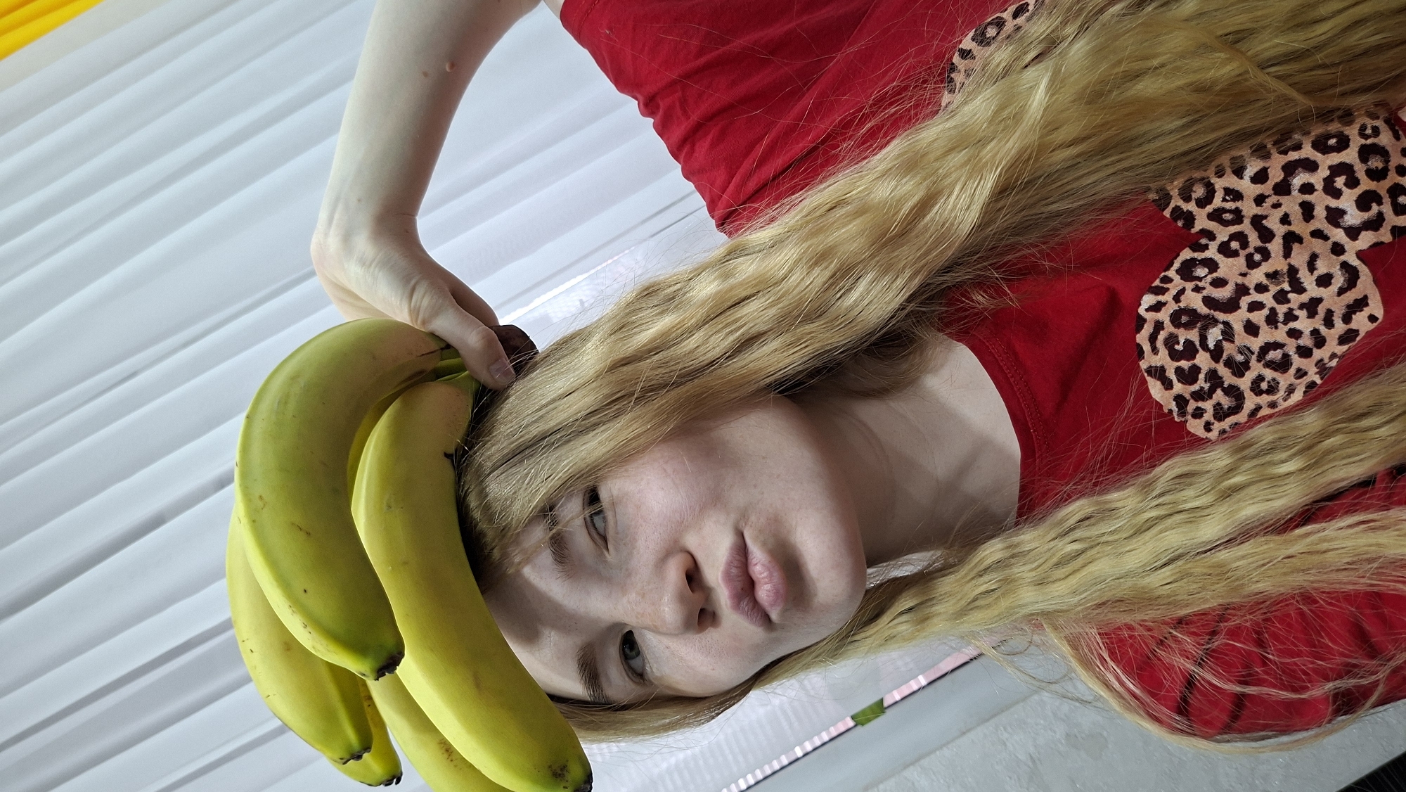 An indoor image depicts a person with long, wavy blonde hair and fair skin, captured in a pose where they appear to be lying down. Their face, exhibiting a neutral to subtly pouting expression with a direct gaze, is positioned towards the center-right of the frame. One hand, belonging to the person, holds a bunch of vibrant green bananas near their head, partially nestled in their blonde hair. The person is wearing a bright red top adorned with a distinctive leopard print design on the shoulder or upper chest area. The background features a white, vertically textured surface, possibly a wall with panels or blinds, and a reflective surface below the person, suggesting they are lying on a floor or table. The lighting is bright and even, consistent with an indoor environment. There is no discernible text. This scene takes place in Chaikovsky, Russia.