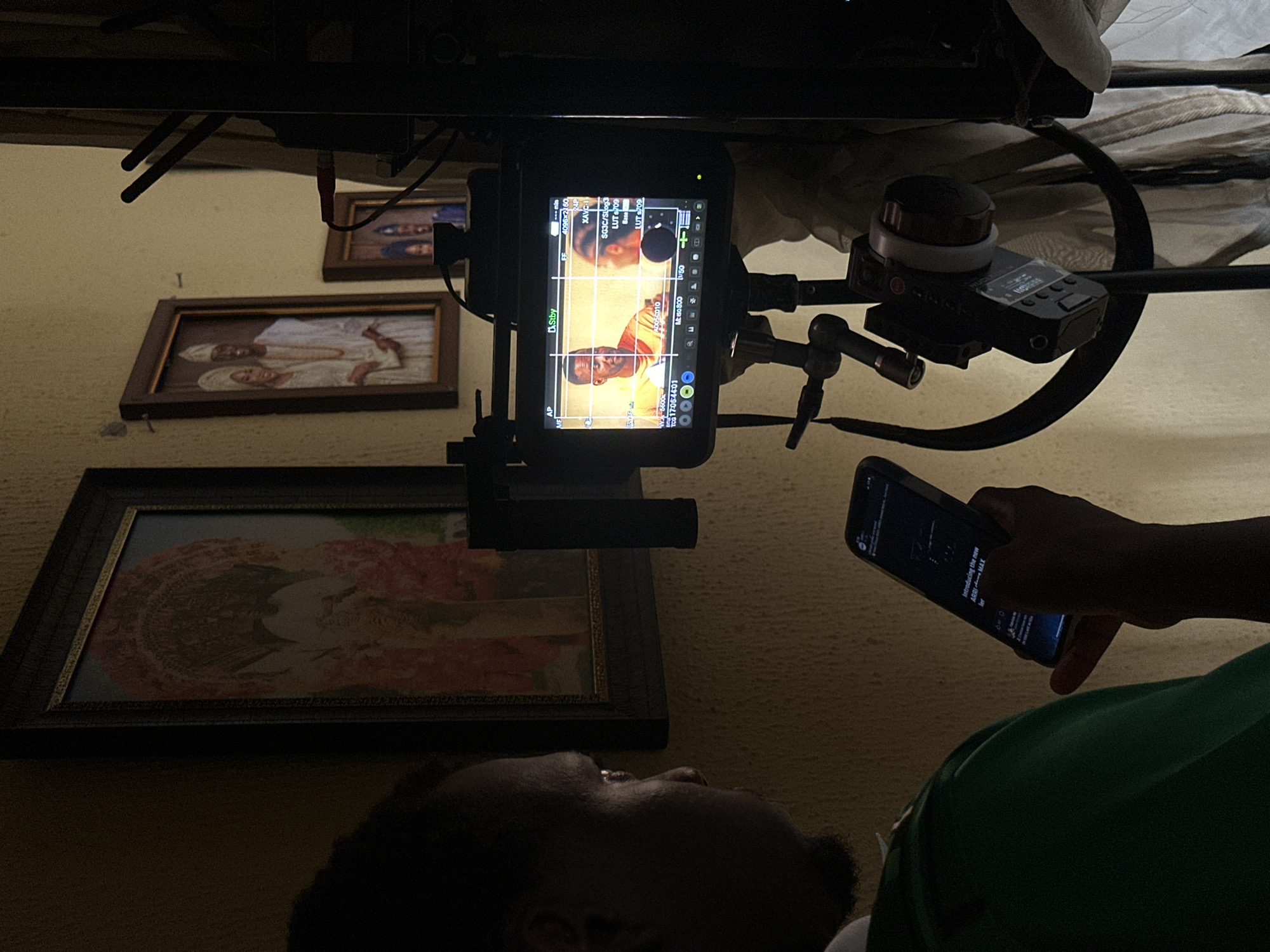 The image captures an indoor scene from a low-angle, upward perspective, seemingly rotated, depicting a person observing a production monitor and a smartphone. The setting is likely a film or video production environment in Lagos, Nigeria.
In the bottom left, a person's head and shoulder are visible, mostly in shadow, looking upwards. They wear a green shirt. On the right, a hand holds a smartphone, its illuminated screen displaying content.
Central to the image is a production monitor mounted on a camera rig, displaying footage of a man with dark skin wearing an orange shirt, shown in a close-up. The monitor screen is rich with technical overlays including "4096x2160" resolution, "24P" frame rate, "XAVC-I" codec, "SG3C/SLog3" log profile, and "LUT S709". Other visible details include "Stby" (Standby), "1/50" shutter speed, "M:50 800", "W-5600", and "TCG 17:06:44:01". Above the monitor, part of a focus pulling system with a large wheel is attached to the rigging.
On the wall behind the equipment, three framed pictures are visible. One prominent picture features two individuals in traditional white attire. Another, larger frame below it, displays a portrait of a person in elaborate traditional clothing with a large headpiece.
The overall lighting is dim, with the monitor and smartphone screens providing significant illumination. The smartphone screen displays text that reads "Introducing the new ARRIMAX" and "600". The Arabic word "محدد" (selected) is also visible on the monitor.