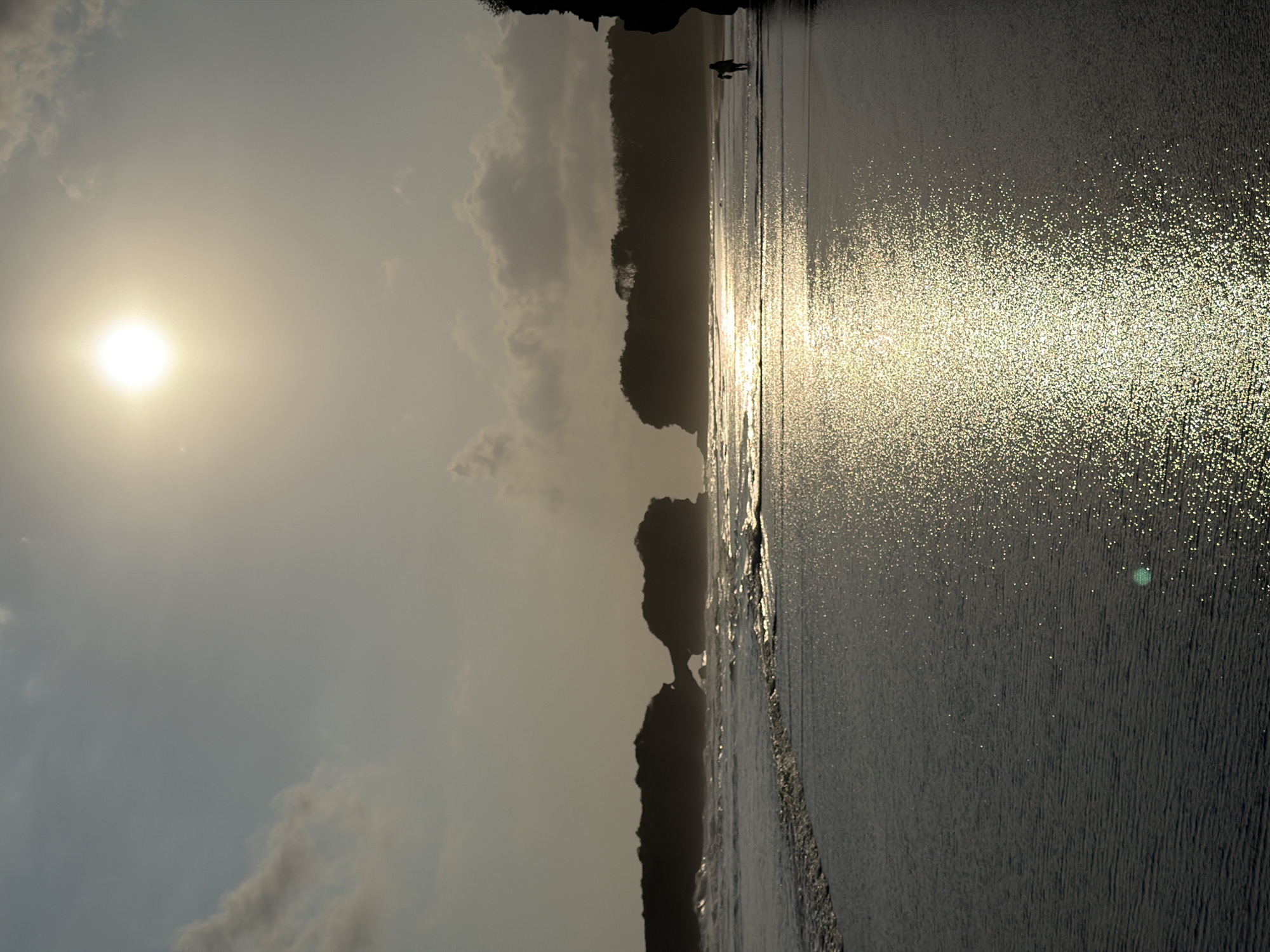 A full shot captures a beach scene in Selemadeg, Indonesia, at sunset. The sun, a bright orb, illuminates the sky and casts a shimmering reflection on the wet sand. In the distance, silhouetted against the light, are rock formations and a treeline, with waves crashing against the shore. A lone figure walks along the beach, heading towards the right of the frame. The atmosphere is serene, with a sense of calm pervading the seascape.
