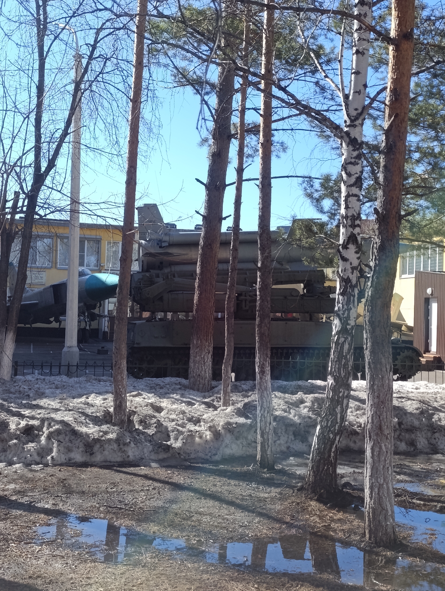 A photograph captures a scene outside, likely in Krasnoyarsk, Russia, displaying a military vehicle and aircraft behind a row of slender trees, with a building in the background. The vehicle, appearing to be a missile launcher on tracks, is parked behind a low, dark metal fence, beside an older gray jet. The background building is a yellow, two-story structure with dark-framed windows. Patches of melting snow remain in the foreground. The ground shows signs of thawing. Puddles reflect the sky and trees. The trees are bare or sparsely foliaged, suggesting early spring or late autumn. The sky is blue with no clouds.