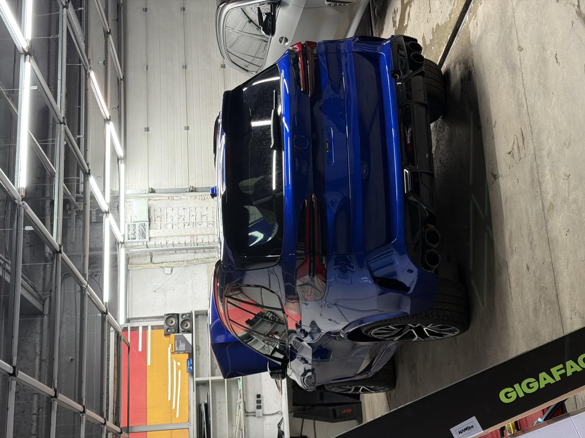 The image shows the rear view of a blue car, potentially a hatchback or SUV, in a brightly lit indoor space that appears to be a car repair shop or detailing facility. The car is parked on a concrete floor, and there is another light-colored car parked nearby. The background includes scaffolding and industrial lighting fixtures on the left side of the frame. A sign partially visible at the bottom reads "GIGAFACT..." and includes a small sticker that reads "NANOLink". The image does not include any people.
There is no information to conclude that the location is in Krasnoyarsk, Russia.