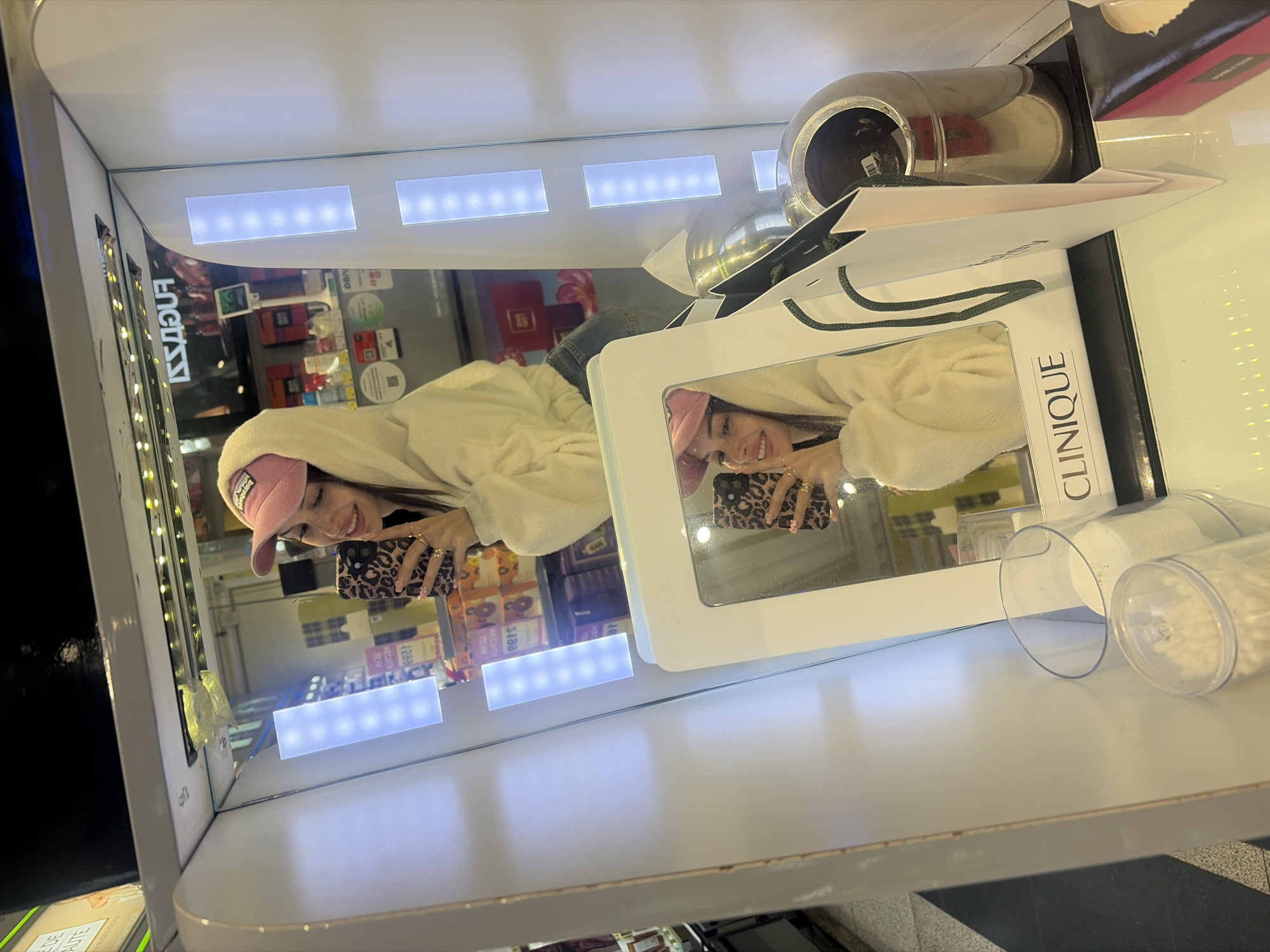 The image depicts a young woman with long, dark hair, wearing a pink baseball cap and a cream-colored hooded sweatshirt. She is holding a smartphone with a leopard print case and taking a selfie in a brightly lit retail display, likely in a department store or cosmetics shop. The display features vertical LED lights and a mirror. In the reflection of the mirror, the woman's face is visible, and she is smiling. Below the mirror, the "CLINIQUE" logo is visible on a product display. To the right of the display, a silver-colored trash receptacle and a white paper shopping bag are present. In the background, shelves stocked with various products are visible, suggesting a retail environment. The floor has a black and white checkered pattern. The overall scene suggests a casual shopping experience. Without further information, it is not possible to determine the specific location in Saint Petersburg, Russia.