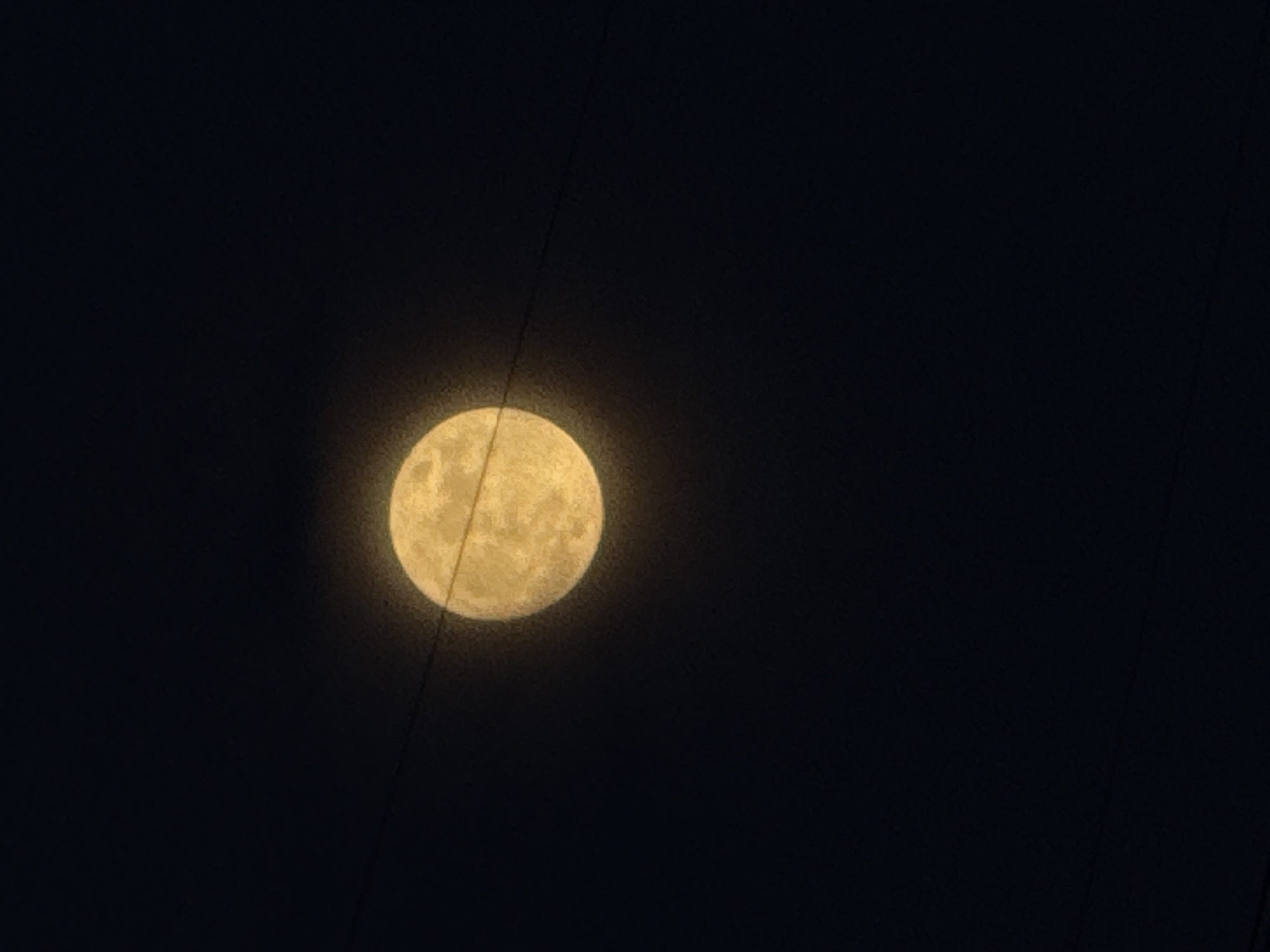 The image presents a striking nocturnal scene featuring a luminous, yellowish full moon against a deep black sky in Chaikovsky, Russia. The moon, the primary subject, occupies the central left portion of the frame, showcasing visible surface details and craters. Its light radiates softly, creating a subtle glow around its edges.
Two thin, dark, linear objects, likely overhead power lines or utility cables, diagonally cross the composition. One prominent line passes over the top-left quadrant of the moon, extending upwards into the dark sky. Another, slightly less distinct line, is visible intersecting the lower-right edge of the moon and extending downwards. These lines partially obscure the moon but also add a contextual element, suggesting an urban or suburban setting.
The time of day is clearly night, indicated by the dark sky and the illuminated moon. The weather appears clear, with no clouds or atmospheric haze obstructing the view of the moon. No discernible human activity or specific event is taking place, beyond the implied act of observing the moon. There is no visible text within the image. The overall impression is one of peaceful observation, contrasting the natural beauty of the moon with subtle signs of human infrastructure.