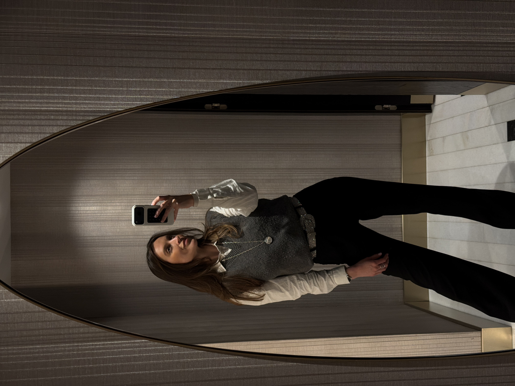 The image shows a woman reflected in a mirror, holding a smartphone and seemingly taking a selfie. She is wearing black pants, a white collared shirt, and a grey vest, with a silver belt and necklace. The mirror has an oval shape with a dark trim. The background visible in the reflection includes textured wallpaper with horizontal lines and a light-colored wall with rectangular patterns. Based on the architecture and interior design, it's plausible the scene is located in a modern hotel or upscale apartment complex in Budva, Montenegro, but without specific landmarks, a precise location is impossible to determine.