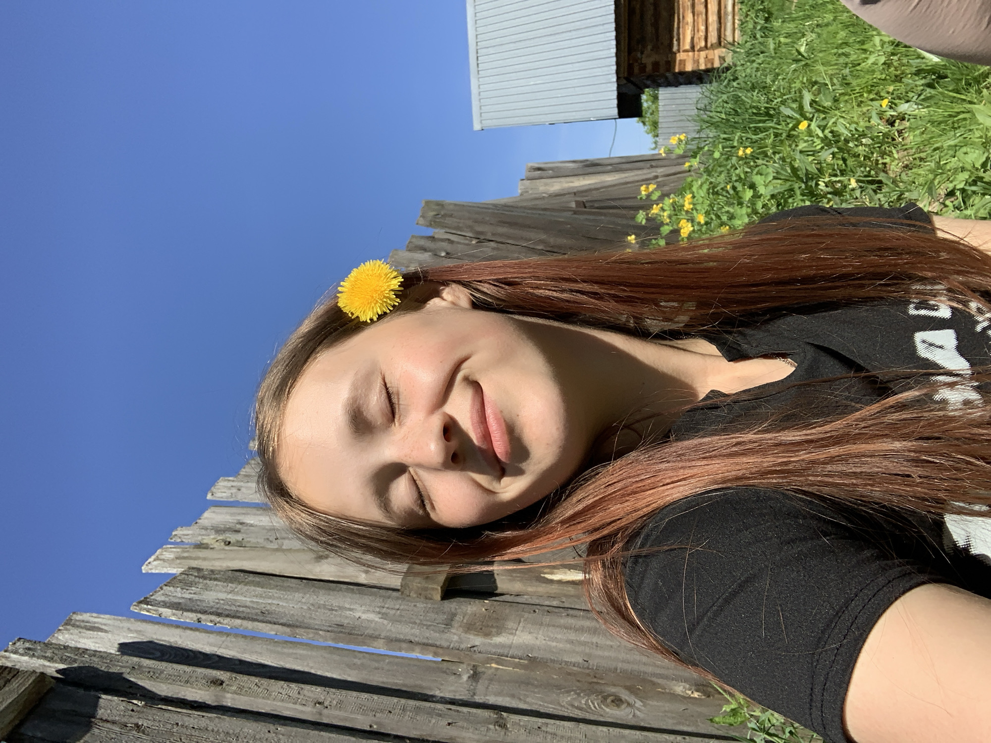 A young woman lies on a weathered wooden fence. Her eyes are closed, and she appears to be smiling gently. A bright yellow flower is tucked into her hair above her ear. She is wearing a black t-shirt. A wooden structure is visible behind the fence, along with patches of green vegetation and small yellow flowers. The sky is a clear, bright blue. The photograph was taken in daylight. I don't have enough information to identify the location of this image in the city of Фоки, Russia.