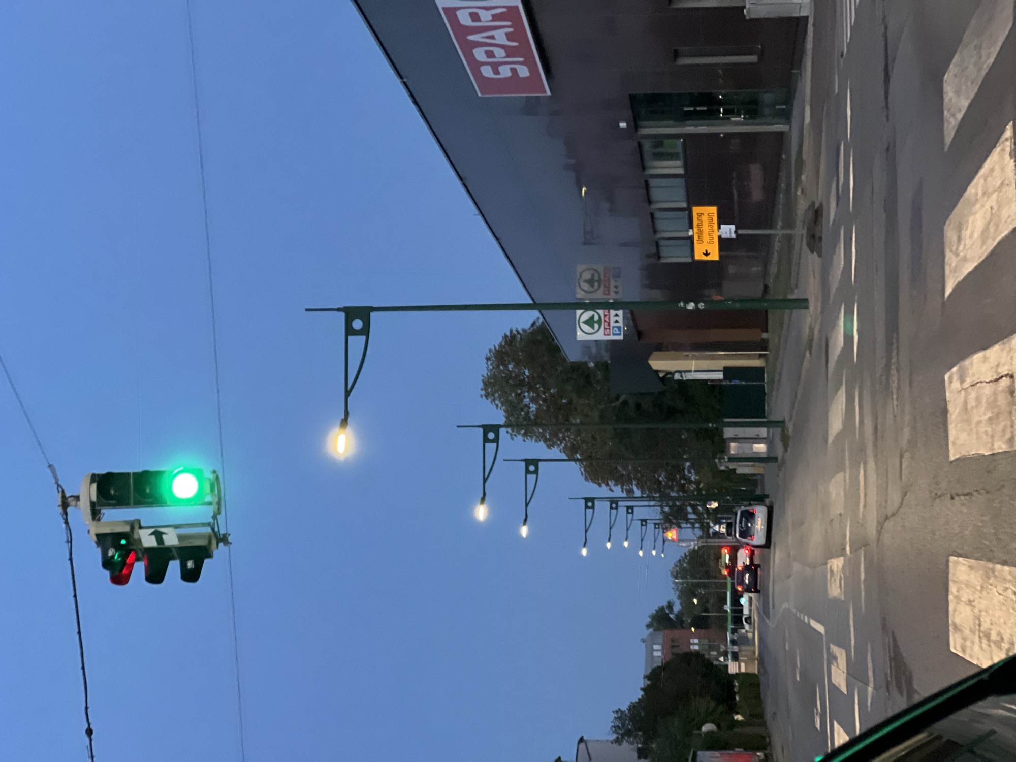 The image depicts a street scene in Mödling, Austria, during twilight. A traffic light with a green signal is prominent in the upper left quadrant. In the foreground, a pedestrian crossing is visible on the asphalt road. To the right, a dark-colored building with a red "SPAR" logo dominates the scene. A row of streetlights with illuminated bulbs lines the left side of the road, extending into the distance. Several cars and a bus are on the road. A yellow directional sign indicating "Umleitung" (detour) with an arrow pointing left is posted near the building. The sky is a deep blue, characteristic of dusk.