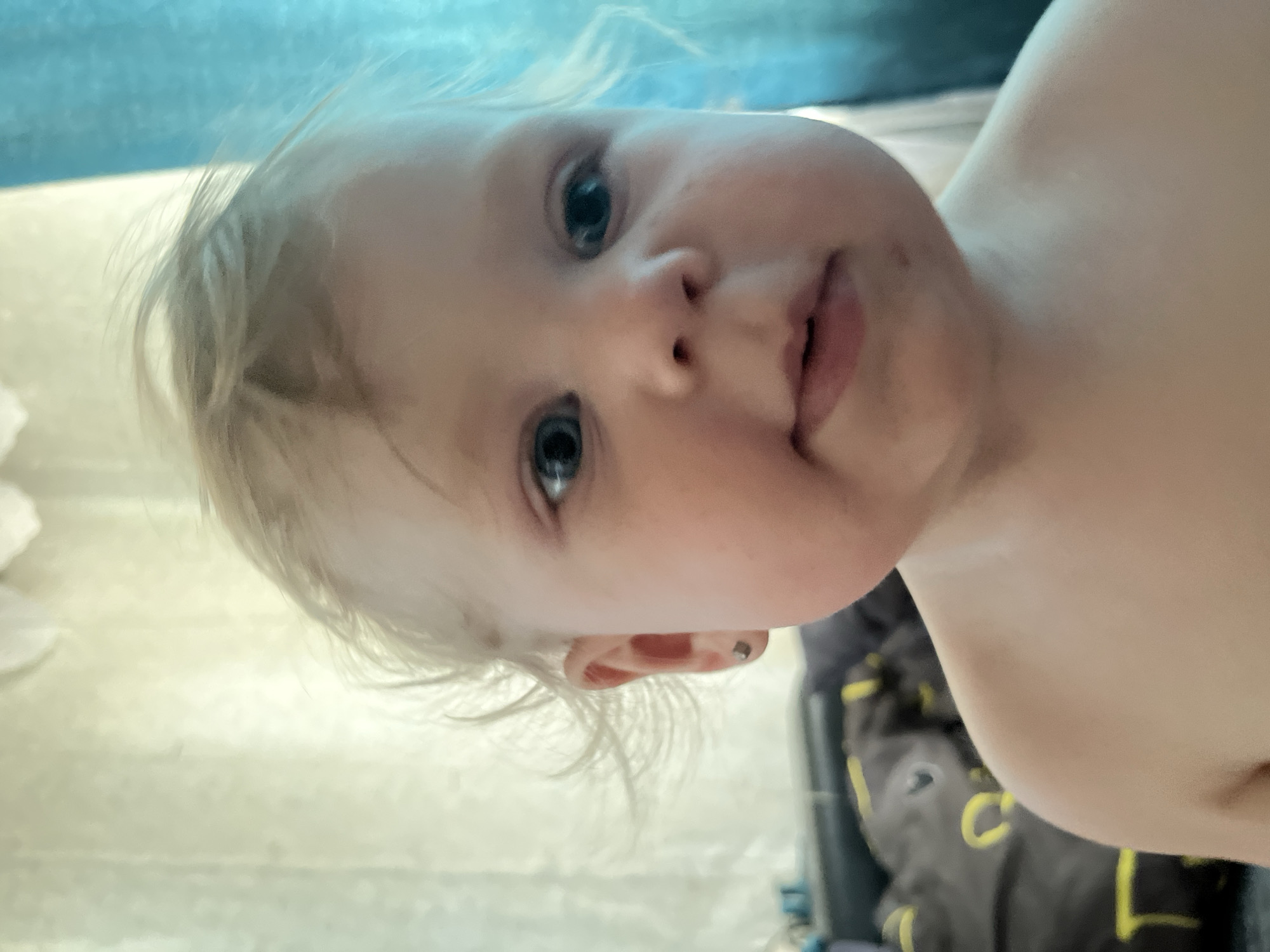 The image is a close-up, head-and-shoulders portrait of a young, fair-skinned child with blonde hair. The child is looking directly at the camera with wide, blue eyes. Their lips are slightly parted, and there are subtle smudges of dark color on their chin and cheek. A small, dark stud earring is visible in the child's left ear. The child's skin appears smooth and rosy.
The background is out of focus. To the left, a blurred object with a dark base and yellow accents is visible, possibly a piece of furniture or bedding. Behind the child, there are curtains, one appearing to be a light, textured fabric and the other a bright blue. The lighting suggests an indoor setting, possibly a room with natural light coming from behind. There is no discernible information in the image that would allow for determination of location within the city of Фоки, Russia.