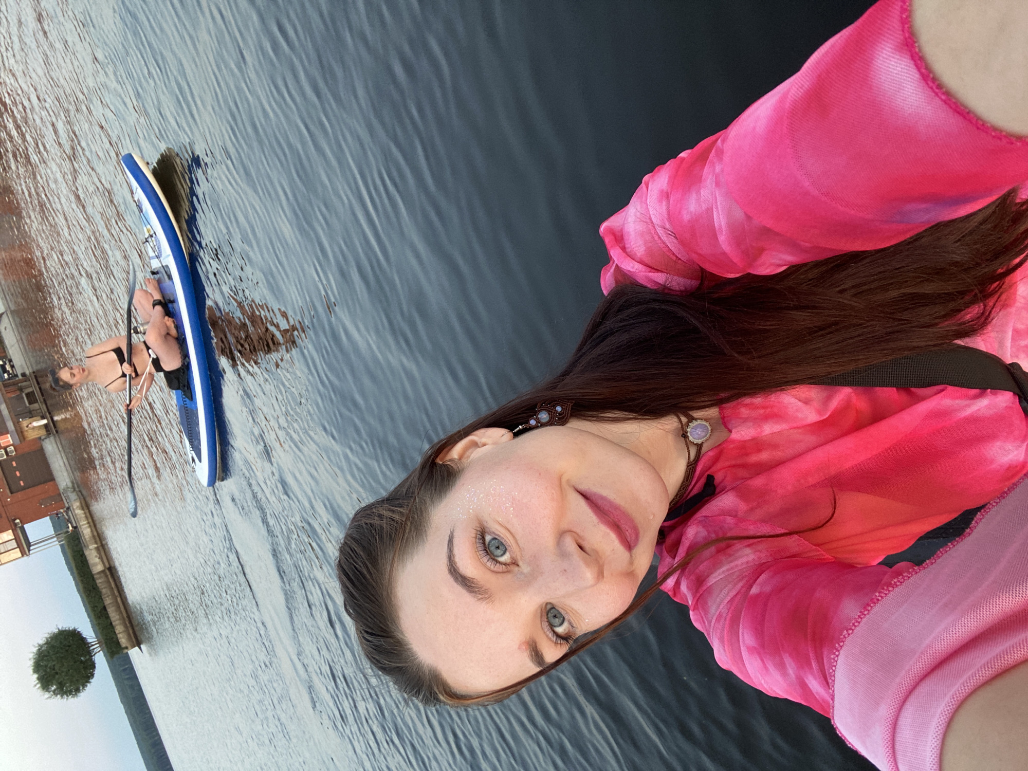 In Chaikovsky, Russia, on a body of water, two individuals are engaged in paddleboarding. In the foreground, a woman with long, dark hair and blue eyes is taking a selfie, smiling and wearing a bright pink, tie-dye shirt and a necklace. She has glitter on her cheek. In the background, another woman with dark hair pulled back is sitting on a blue and white paddleboard, holding a paddle and looking to the side. She is wearing a black bikini. The water is rippled, and a building with a red brick facade and a single tree are visible on the shore in the distance. The sky is overcast and appears to be dusk.