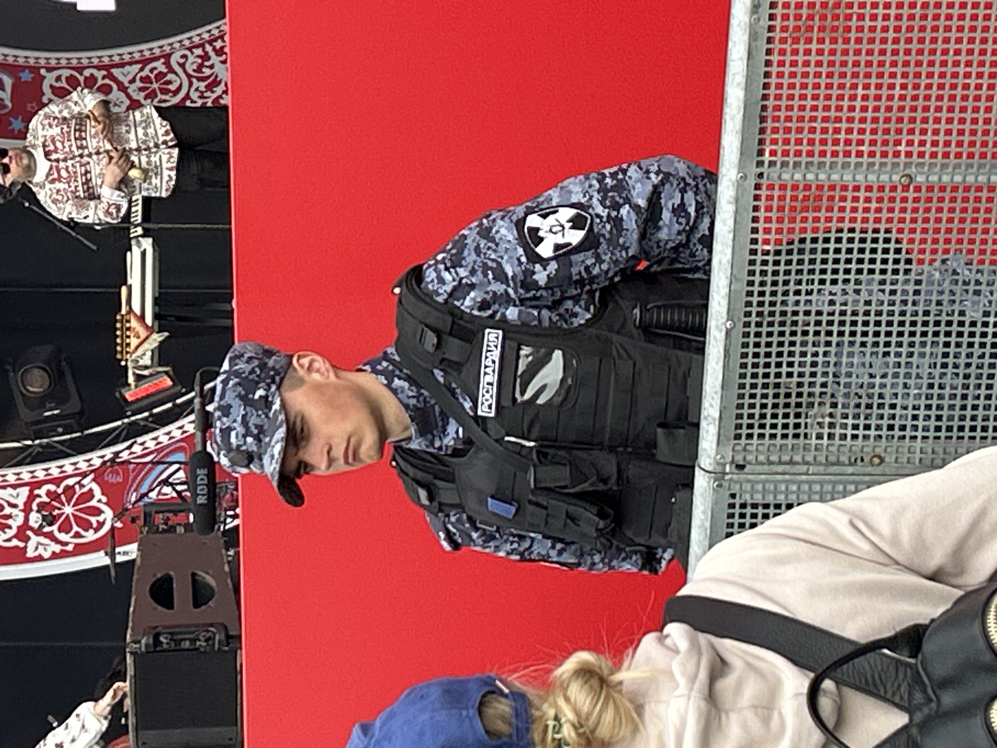 A man in a grey camouflage uniform and a black vest with "РОСГВАРДИЯ" (Rosgvardiya) on it, standing in front of a red backdrop with a metal fence to his right. The patch on his left shoulder features a black symbol with an atom-like design. Other individuals and objects are partially visible in the foreground. The location is likely Moscow, Russia due to the Rosgvardiya insignia.