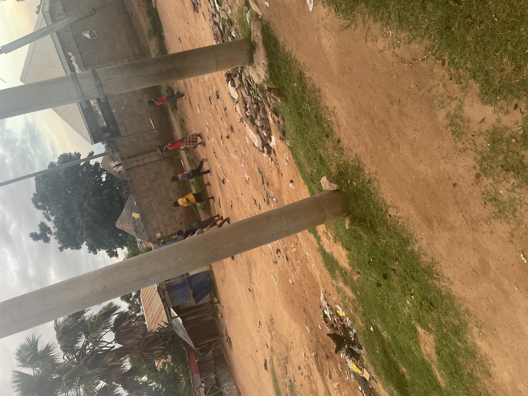 The image depicts a street scene in Jalingo, Nigeria. In the foreground, a dirt road with patches of grass is visible. Two utility poles stand prominently in the middle ground. To the left, a pile of garbage with a chicken scavenging is present. Several children, appearing to be walking or playing, are scattered across the middle and background. They are dressed in casual clothing, with one child wearing a red and white checkered skirt. In the background, a few buildings with corrugated metal roofs and brick walls are visible, along with some palm trees and other vegetation. The sky is overcast.