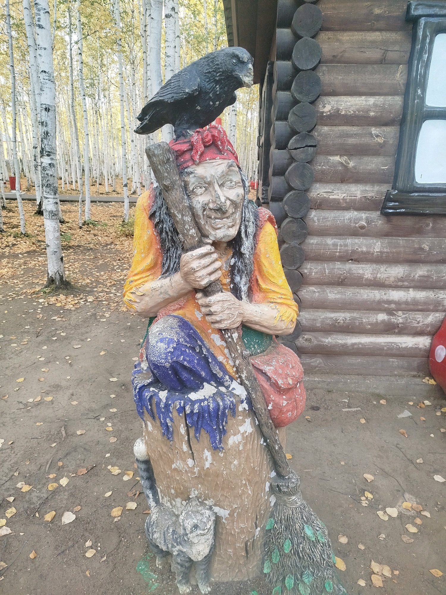 This is a medium-full shot of a carved, weathered statue of a witch and a crow, positioned outdoors in front of a log cabin. The witch is seated on a stump, holding a broomstick and a wooden staff. She has a wrinkled, aged face with dark braids and is wearing a yellow top, blue pants, and a red headscarf. A red satchel hangs from her shoulder. A black crow is perched on top of her head. To the left of the witch is a small, carved grey cat. The ground around the statue is covered with fallen yellow and brown leaves. In the background, a dense forest of white-barked birch trees can be seen. The log cabin behind the statue has dark logs and a window with a black frame. The overall atmosphere is autumnal. This statue appears to be located in an outdoor park or garden, possibly a tourist attraction, in the city of Mirny, Russia, given the presence of birch trees, characteristic of the region.