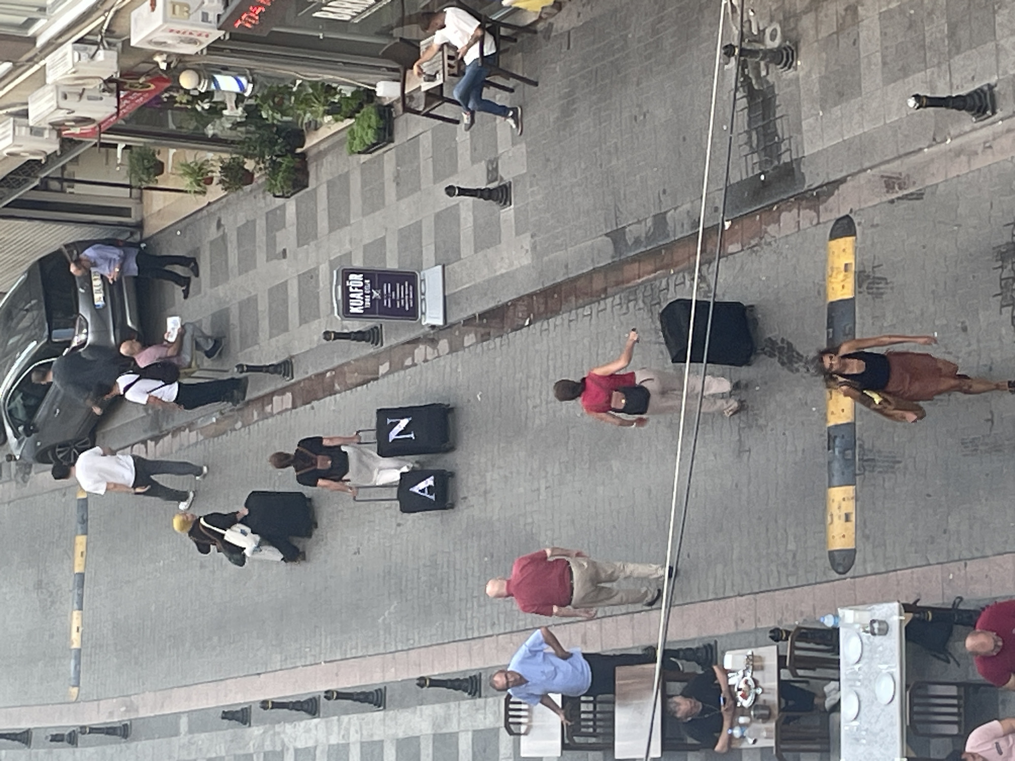 This high-angle, full-color photograph depicts a street scene in Kadıköy, Turkey, likely on a cobblestone street with gray and beige checkered pavement sections. Several individuals are visible, some walking and others seated at outdoor tables.
In the upper portion of the image, a black car is parked on the street. A man in a white t-shirt and dark pants walks away from the camera. To his right, a person with blond hair and a yellow beanie is seen from behind, pulling a large black suitcase. Further down the street, a person in a white t-shirt and white pants is walking away, pulling two black suitcases, one labeled with the letter "A" and the other with "N." Another person in a red top and khaki pants is walking away, pulling a black suitcase.
In the middle ground, a man in a light blue short-sleeved shirt and dark pants stands with his hand on his hip, facing the camera. To his right, a man in a red short-sleeved shirt and light brown pants walks away from the camera. Further right, a man in a white t-shirt and blue jeans sits at a small outdoor table, holding a phone and a cup. In front of him, a barber pole with red, white, and blue stripes stands near the entrance of a shop labeled "KUAFÖR." Plants are visible in pots outside the shop.
In the lower portion of the image, several people are seated at outdoor tables. One man, wearing a black t-shirt, is seated at a table with food and drinks in front of him. Another table is set with plates. A woman in a black tank top and a brown skirt walks away from the camera, carrying a brown purse over her shoulder.
The environment includes lampposts along the side of the street, a speed bump on the road, and various outdoor seating areas. Electrical wires are visible crossing the scene.