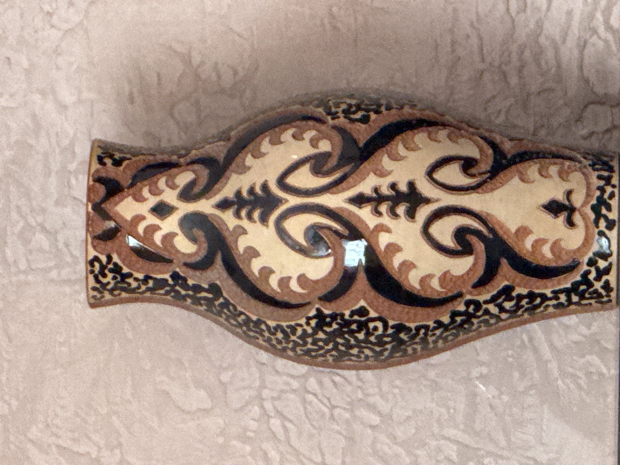 A close-up, eye-level, full-shot photograph depicts a ceramic vase with an ornate, curvilinear pattern. The vase is predominantly tan with intricate detailing in brown and black. The pattern features scrolling motifs and stylized floral elements. The background is a textured, light-colored surface, possibly a wall or fabric, which appears to be a light pinkish-beige with subtle shadowing that suggests folds or undulations. The lighting is diffuse, highlighting the texture of the background and the glossy finish of the vase. There are no people or other objects visible in the frame. The scene is static and appears to be a studio shot or an indoor display of the vase. The location is not identifiable from the image itself.