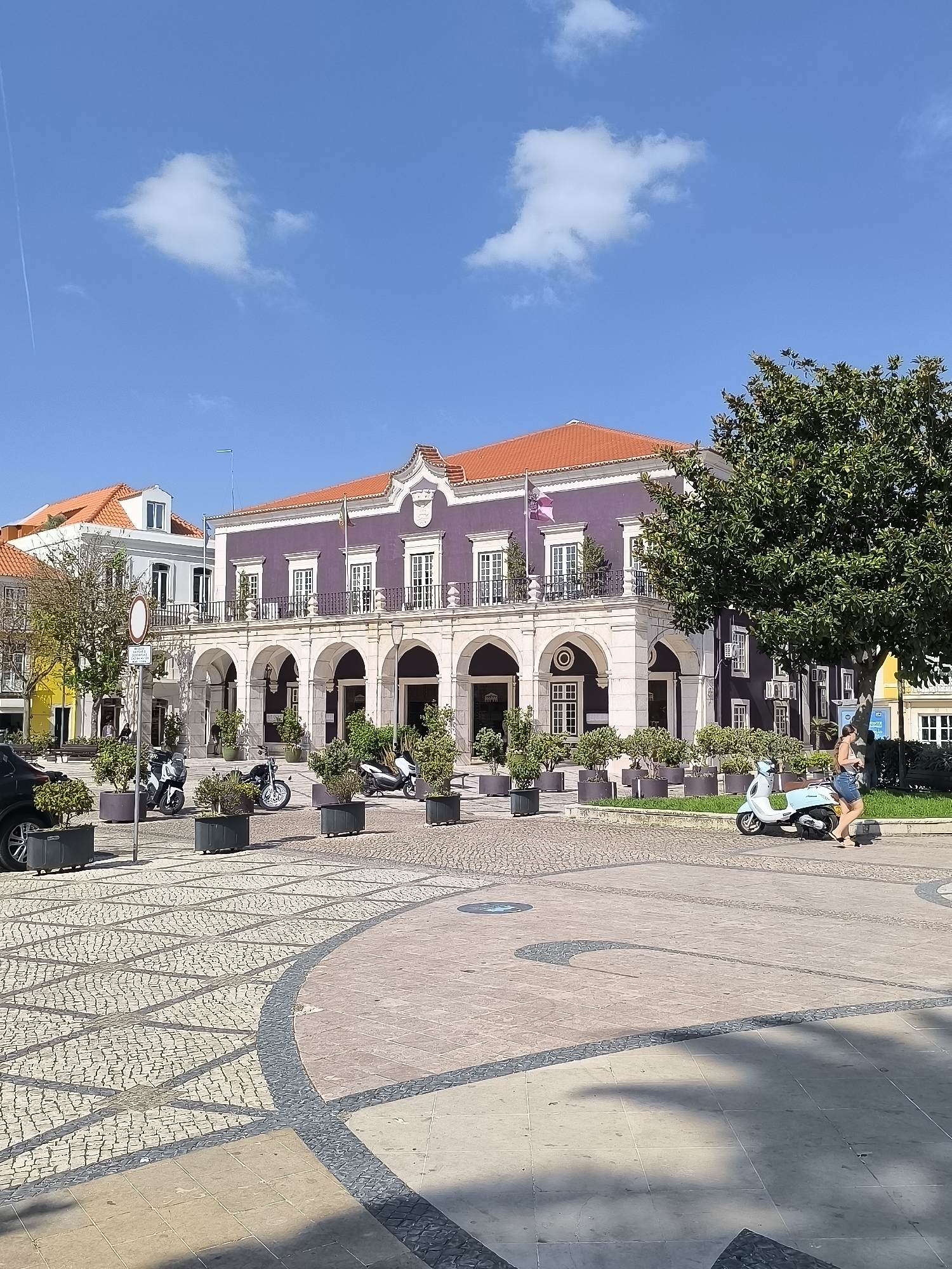 This is a medium shot of a picturesque town square on a sunny day. A prominent building with a purple facade and a terracotta roof dominates the background. The building features a covered arcade with arched entrances. In front of the building, several potted plants add greenery to the scene.
To the left of the main building, another building with a white facade and a similar terracotta roof is partially visible. The foreground of the image is a paved square with intricate geometric patterns made of cobblestones and larger tiles.
Several objects are present in the square: a black car on the far left, three motorcycles parked in front of the potted plants, and a light blue scooter. A woman in a denim skirt and a dark top is walking past the scooter.
The sky is a clear, vibrant blue with a few wispy white clouds, and a contrail from an airplane streaks across the upper left corner. The sunlight casts distinct shadows on the square, suggesting it is midday. The overall atmosphere is calm and pleasant, indicating a leisurely day in what appears to be a historic European town.
There is a circular traffic sign with a red border and a white interior on a pole to the left of the main building, and some text on a blue sign attached to the building on the right. No other visible text is legible. Given the location context of Setúbal, Portugal, this scene likely depicts a public space in that city.