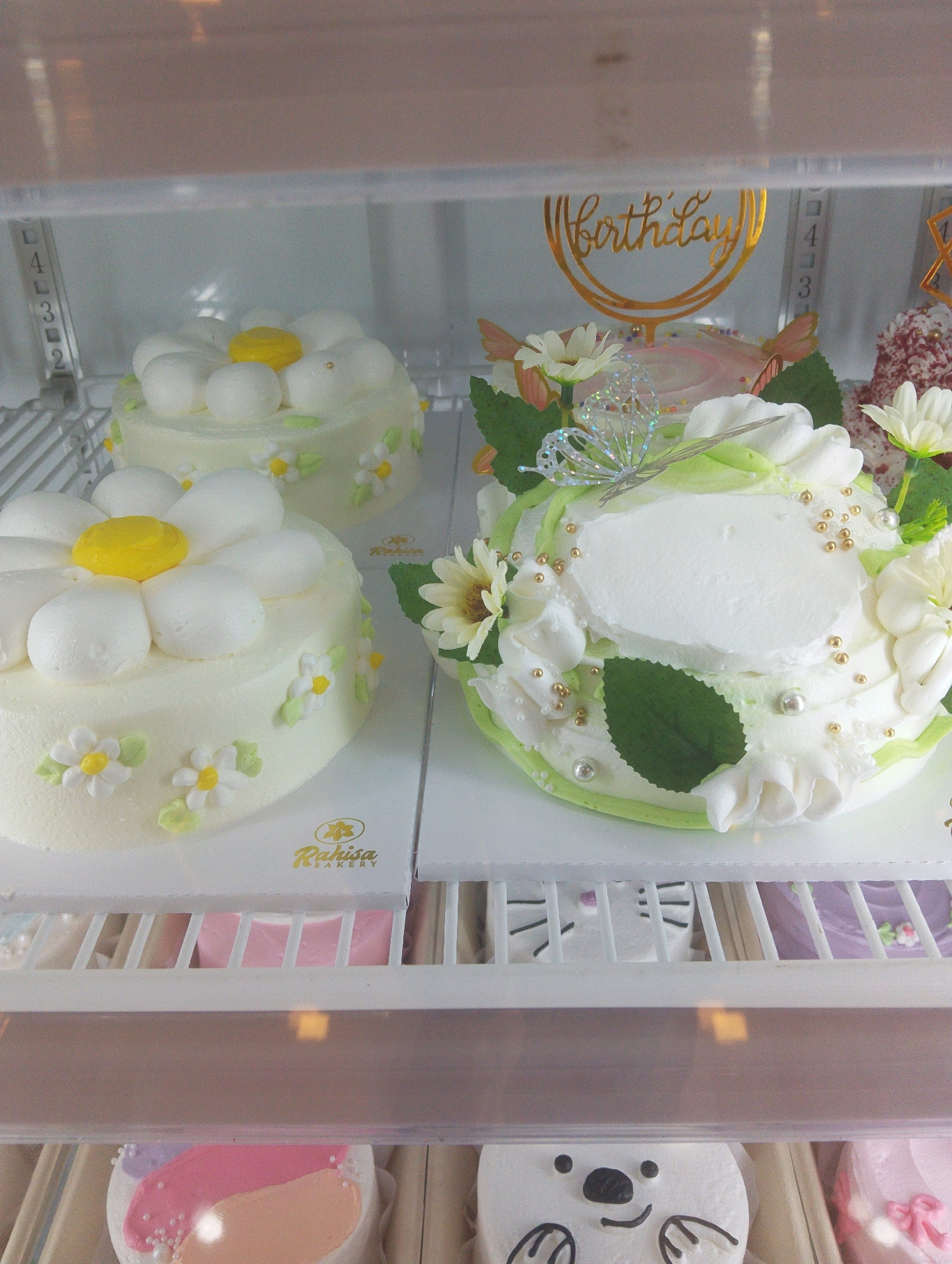 A close-up, eye-level interior shot shows three cakes displayed on shelves in a refrigerated case. The cakes are decorated with white frosting and floral motifs. The cake on the left is round and decorated with white daisy-like frosting flowers with yellow centers and green leaves. The middle cake is also round, with white frosting, green trim, and is adorned with white flowers, green leaves, gold beads, and a sparkly butterfly. A gold "birthday" cake topper is visible behind this cake. The third cake, partially visible on the right, appears to be red and white. The cakes are presented on white cardboard bases. The refrigerator case has clear plastic doors and shelves. A partially visible "Rahisa BAKERY" logo is present on the cake bases. The background includes the interior of a refrigerator with a white rack and a pink object behind the front cakes. The scene does not depict Manaruwi, Indonesia, or any people.