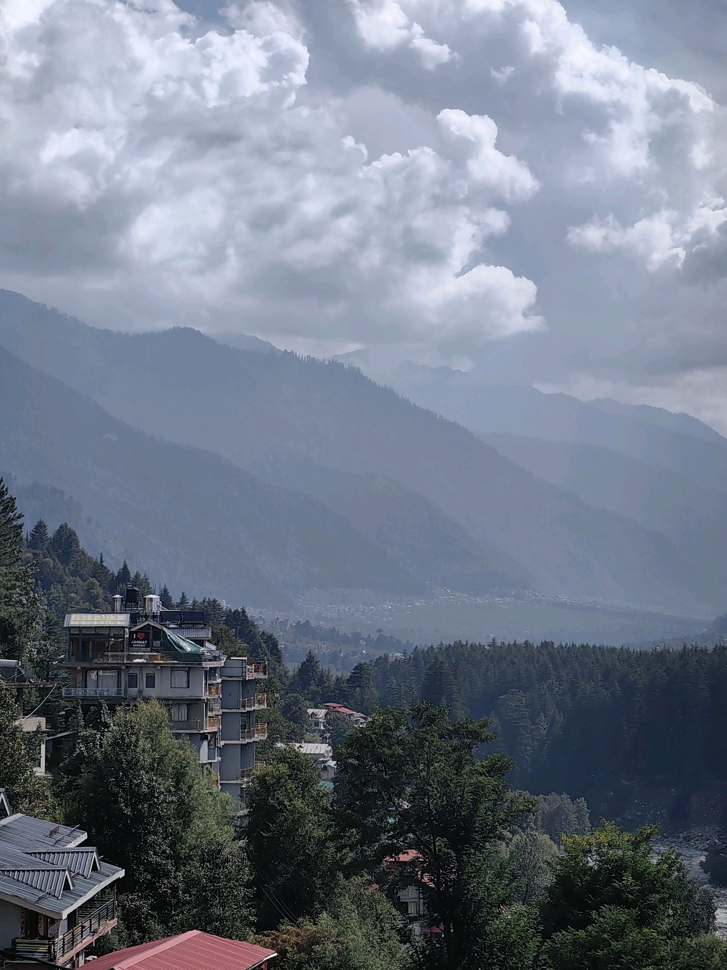 A view from a height shows a mountainous landscape in Vashisht, India, with a cluster of buildings in the foreground and a valley with a distant town and more mountains in the background. The sky is overcast with dramatic clouds. In the immediate foreground, there are several buildings with pitched roofs, some with visible balconies and water tanks. A taller, multi-story hotel with balconies and windows is prominent. The hotel has a sign that reads "I LOVE JANNAT MANALI". Dense green trees surround the buildings. The middle ground features a lush, dark green coniferous forest covering the slopes of the mountains. In the distance, a hazy valley with a small town is visible, with the silhouettes of further, mist-shrouded mountains stretching to the horizon. The overall scene is one of natural beauty and human habitation nestled within a rugged environment.