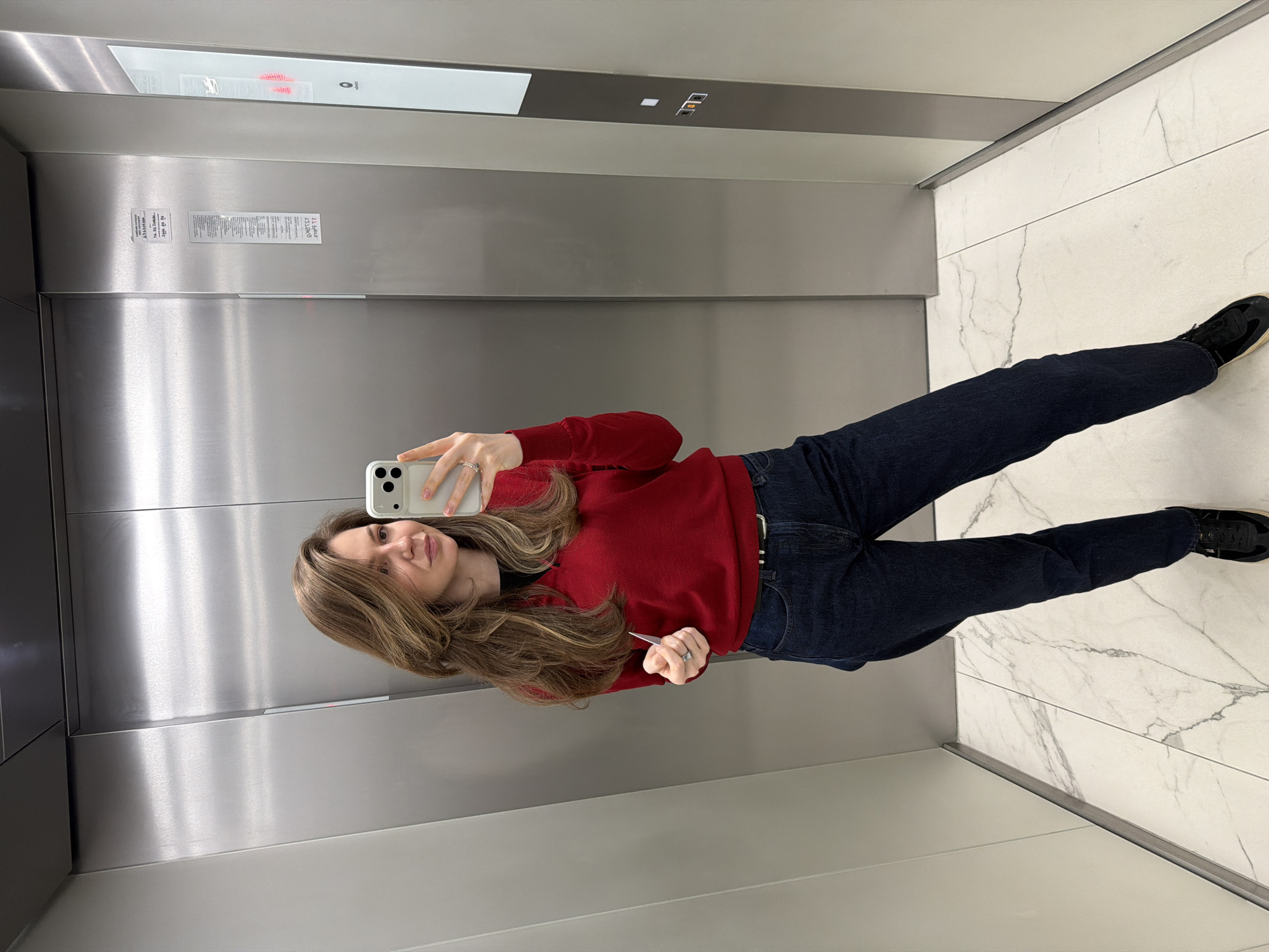 A young woman in dark denim jeans and a red sweater stands in an elevator with brushed metal walls and white marble floors. She is holding a white smartphone in her left hand, taking a selfie. The elevator has signage and a floor indicator panel. While it's impossible to determine the location as Riga, Latvia, without more information, the style of the elevator suggests a modern commercial or residential building.