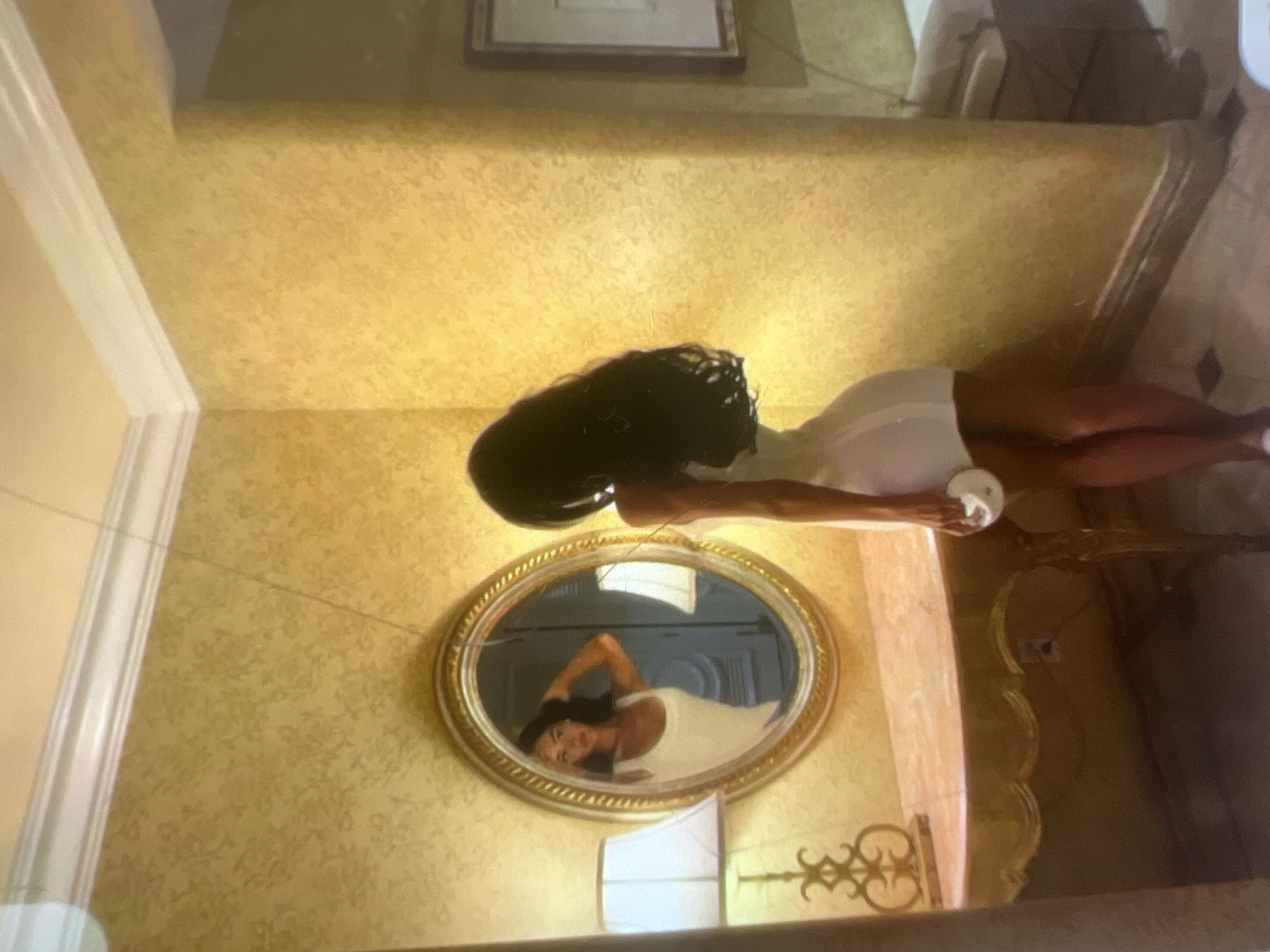 A woman with long, dark hair is standing in a room, facing away from the camera and towards an oval mirror. She is wearing a white, form-fitting dress. In the mirror, her reflection shows her face, and she is touching her hair with her right hand. To the left of the mirror is a lamp with a white shade on an ornate base. The mirror sits on a marble-topped vanity with a gold, ornate base. The walls of the room are covered in textured, yellow wallpaper. To the right of the woman, a framed picture hangs on the wall. The floor appears to be tiled. This scene is located in a room within the city of Kara-Dıykan, Kyrgyzstan.