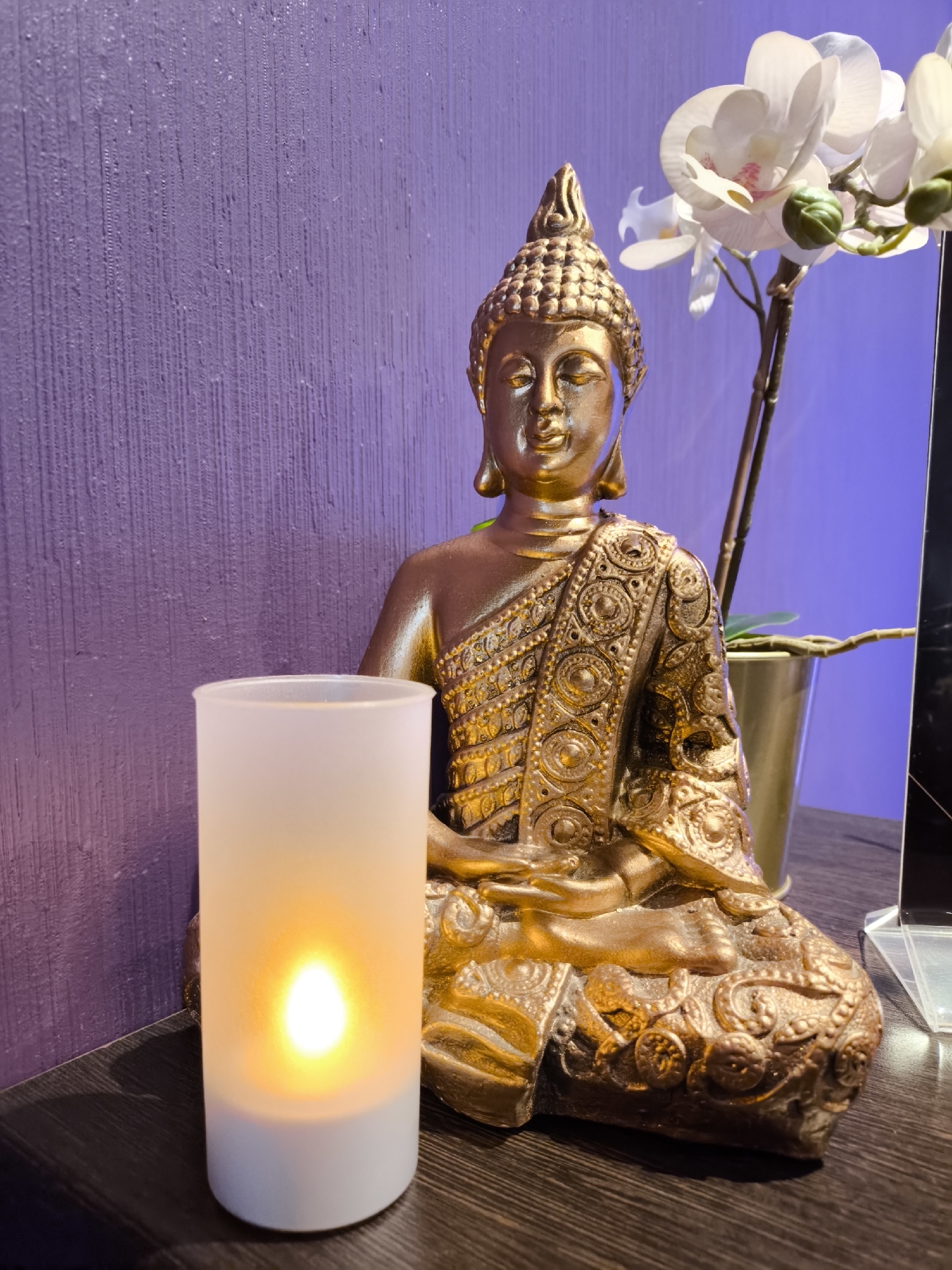 A still life features a golden Buddha statue seated in a meditative pose, juxtaposed with a cylindrical, off-white candle displaying a warm, artificial light. Beside the statue, a golden-colored pot holds a white orchid. The backdrop consists of a textured, solid purple wall that meets the wooden surface on which the items rest. No identifiable location in Moscow, Russia is evident.