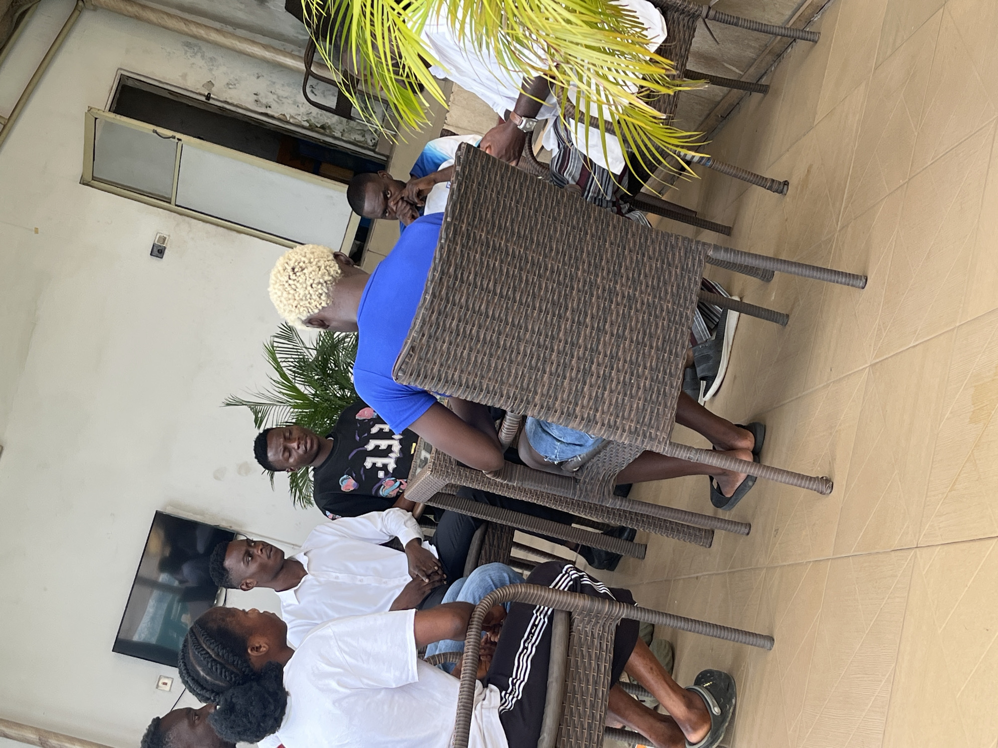 A group of six young adults, appearing to be in their late teens to early twenties, are gathered around a wicker table on an outdoor patio. The patio is paved with light beige square tiles. In the background, a white wall is visible, with a dark flat-screen television mounted on it, along with a light-colored door frame and a partially open door. Lush green foliage is present to the right of the scene, partially obscuring the view.
Three of the individuals are women, and three are men. The woman in the foreground on the left wears a white t-shirt and black pants with white stripes. She has braided hair tied back. A man in a white dress shirt and black pants sits next to her. Another man, wearing a black t-shirt with white lettering, is seated across from them. A man with blond, spiked hair, wearing a bright blue t-shirt, is seated with his back to the camera, facing the table. Next to him, a man in a blue and white t-shirt looks towards the camera, resting his hands on his chin. The sixth individual, a man on the far right, is wearing a white striped shirt and appears to be looking down at the table.
The individuals are seated on wicker chairs around the table. The overall atmosphere suggests a casual gathering or meeting. The scene is well-lit, indicating it is daytime. The architecture and tiling suggest a modern, possibly commercial or residential establishment in Abuja, Nigeria.