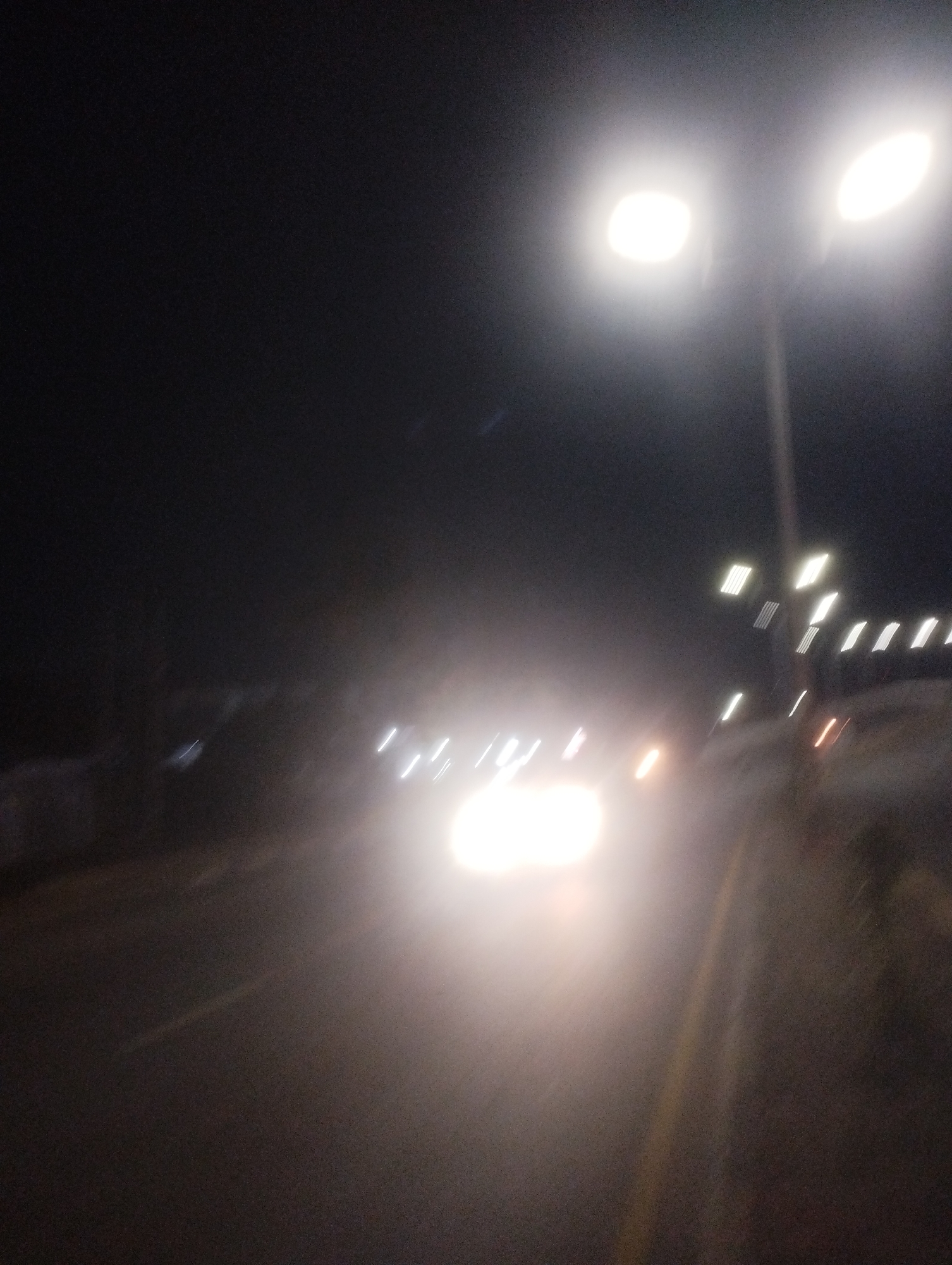 The image depicts a night scene, likely taken from a moving vehicle on a road in Jos, Nigeria. The primary focus is the bright headlights of an oncoming vehicle, which are overexposed and create a strong glare. Above this, two streetlights are visible, also emitting a bright light that causes lens flare. The background is dark, suggesting it is nighttime. The image is blurry due to motion, making it difficult to discern specific details of the surrounding environment, such as buildings, other vehicles, or pedestrians. The road itself appears to be paved, with a yellow line visible on the right side, possibly marking the edge of the lane. The overall impression is one of a dimly lit, urban nighttime setting with significant light sources causing visual disruption.