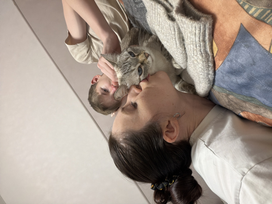 An indoor scene depicts three subjects: two humans and one feline, resting on a bed. In the left foreground, an adult female with dark hair secured in a bun, wearing a light-colored t-shirt and a silver hoop earring, is seen in profile. Her eyes are closed, and her lips are pursed, making direct contact with the left cheek of a cat, indicating a kiss. The cat is a domestic short-haired type with lynx point tabby markings and blue eyes, positioned in the center, looking directly forward. To the right and slightly behind the cat, an adult male with light-colored hair, wearing a light-toned t-shirt, rests his right hand near the cat's head while gazing at it. The subjects are positioned on a bed covered with a patterned duvet featuring blue, brown, and orange geometric designs. A light grey knitted garment lies partially on the bedding. The background consists of a plain, light-colored wall. The scene is located in a residential setting within the city of Perm, Russia.