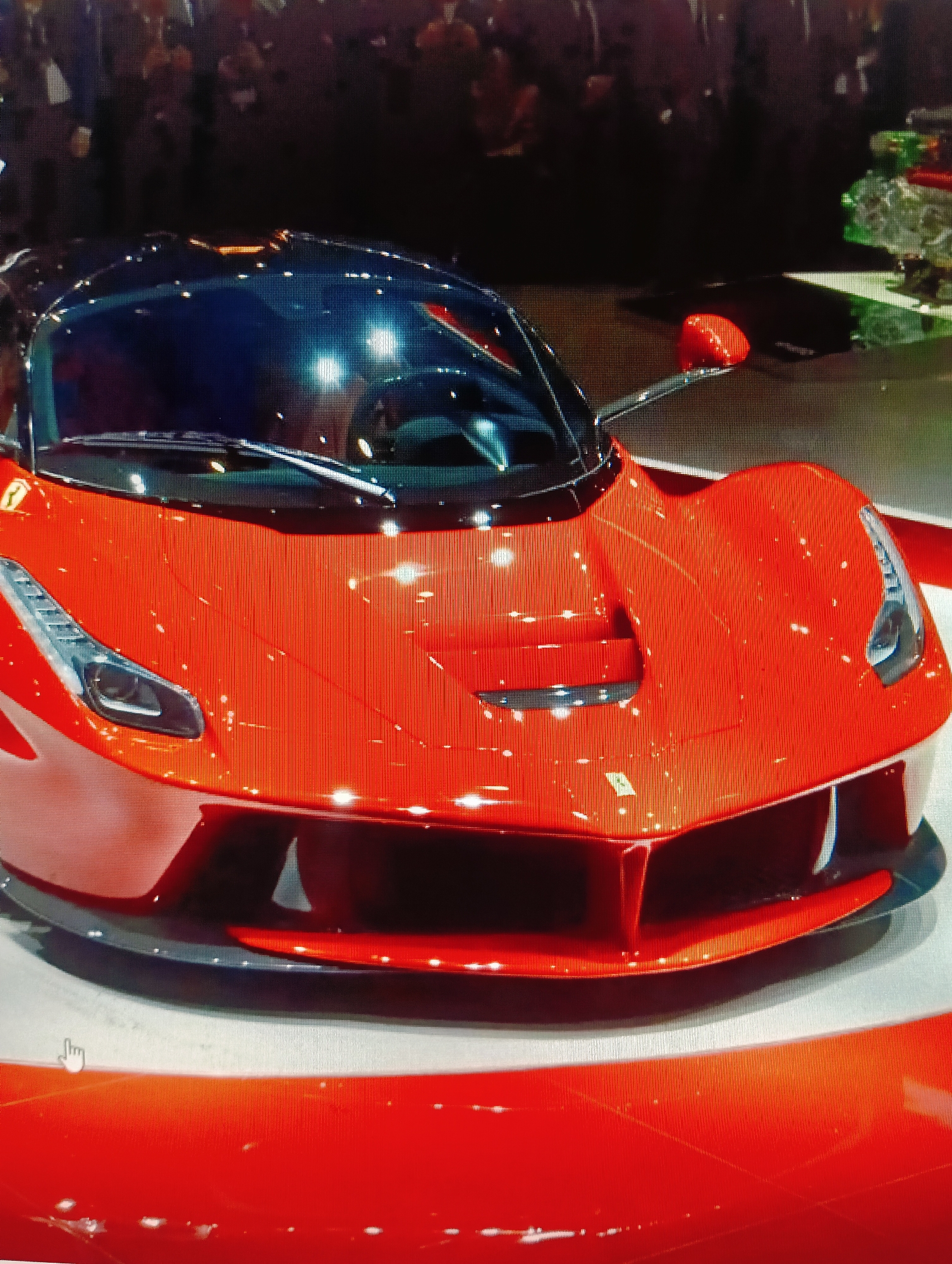 The image shows a red Ferrari supercar on display. The car has a black roof and dark tinted windows. The front of the car features sharp headlights and a prominent grille. The vehicle is positioned on a white platform, and a crowd of people is visible in the background, suggesting a car show or exhibition. There are reflections of lights on the car's glossy surface. The scene appears to be indoors. There is no information about the city of Sluck, Belarus, in the image or its context.