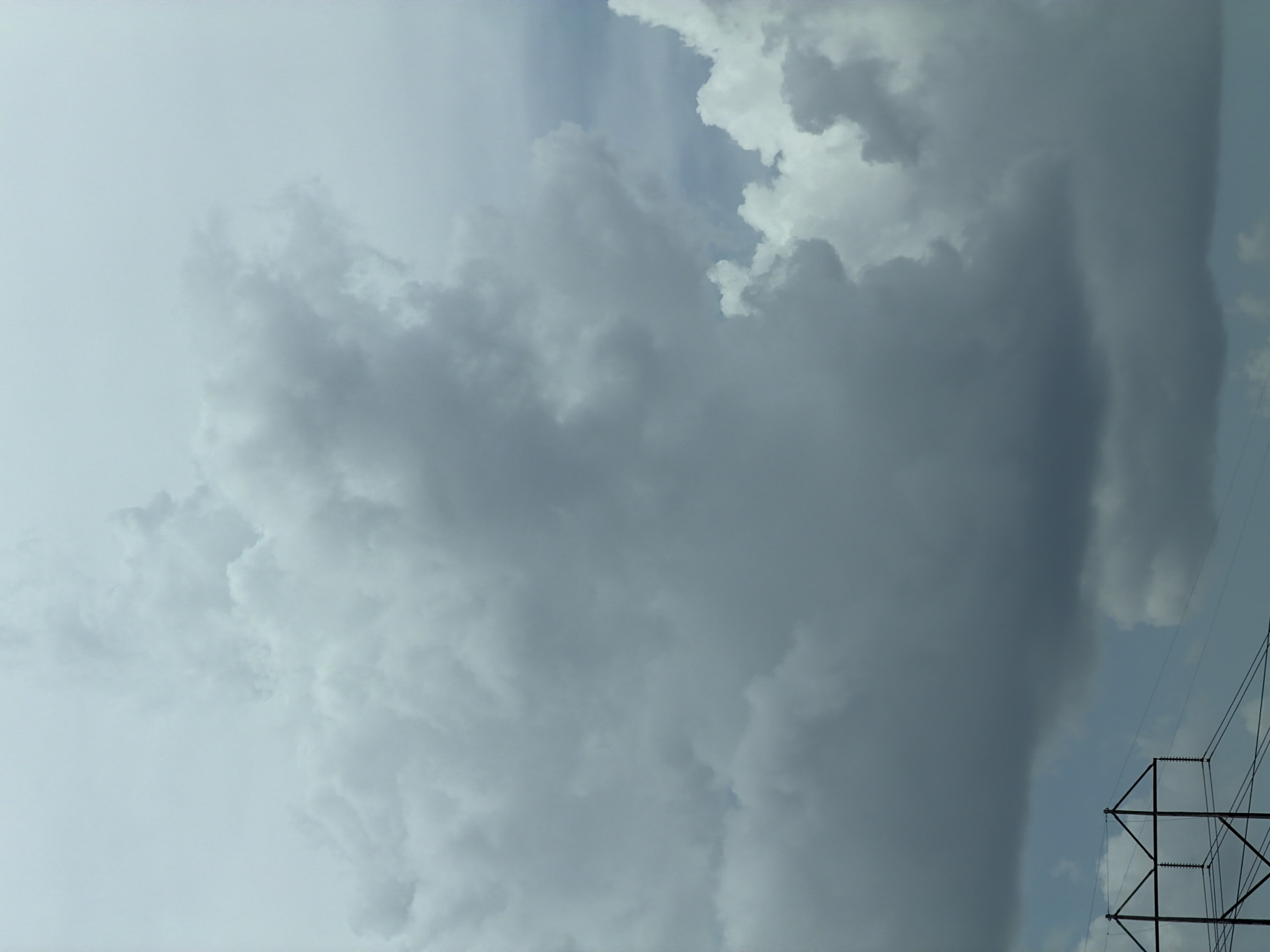 A detailed, matter-of-fact description of the image, focusing on its visual elements, is as follows:
The image depicts a dramatic sky filled with large, dense cumulus clouds. These clouds vary in shade from a bright, almost white hue at their peaks to deep grays and blues in their lower portions, suggesting an impending storm or overcast conditions. The lighting is diffused, creating a soft, somewhat somber atmosphere.
In the lower left quadrant of the image, a portion of a dark, angular electrical pylon or transmission tower is visible. The tower is constructed of dark metal beams forming a geometric, triangular structure at the top, with crossbars supporting what appear to be insulators and cables. Several wires extend from this structure towards the right side of the frame, disappearing into the cloudy sky.
The overall scene captures a natural, yet imposing, display of weather juxtaposed with human infrastructure. There are no people or animals visible in the image. The environment appears to be an outdoor setting, possibly a rural or suburban area given the presence of the power lines and tower. The color palette is dominated by shades of gray, white, and blue, with the dark metal of the tower providing a stark contrast against the sky. The perspective is from below, looking upwards towards the clouds and the upper portion of the tower.