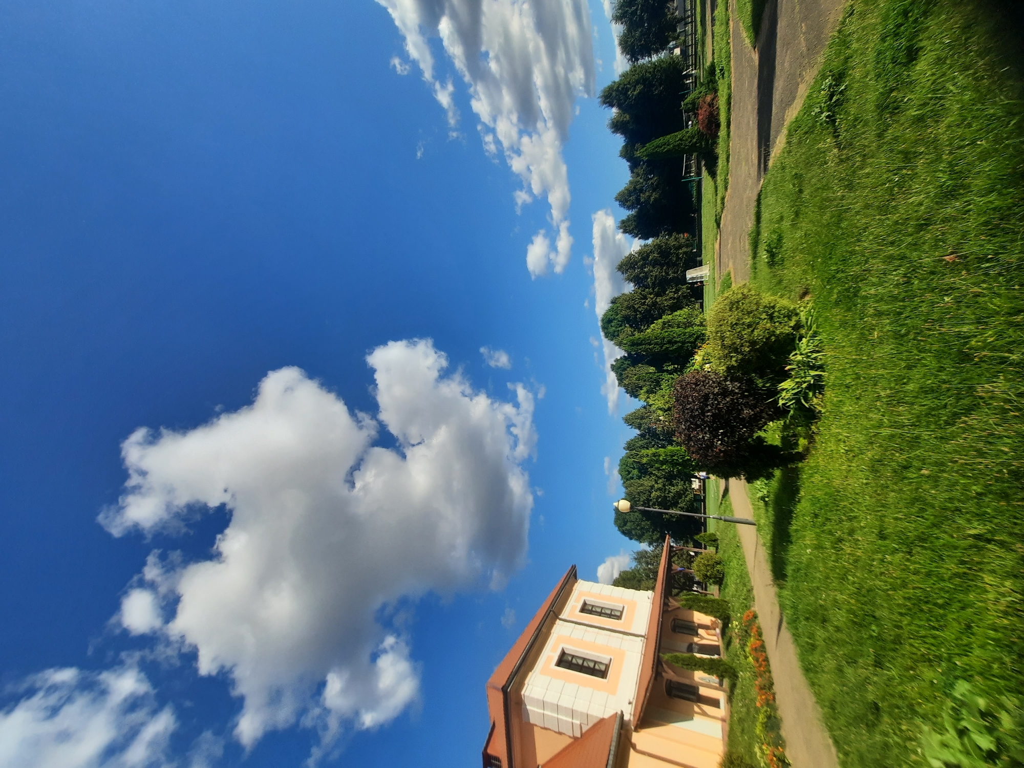 This is an eye-level, wide-angle, outdoor shot taken on a sunny day in Shklov, Belarus. The image features a building on the left, a paved walkway, and a grassy area with trees in the background. The building, with an orange roof and cream-colored walls, has several windows and a covered walkway along its side. A streetlamp stands near the building, and a dark red bush is located on the right side of the walkway. The sky is a vibrant blue with scattered white clouds. In the distance, a line of trees borders a field.