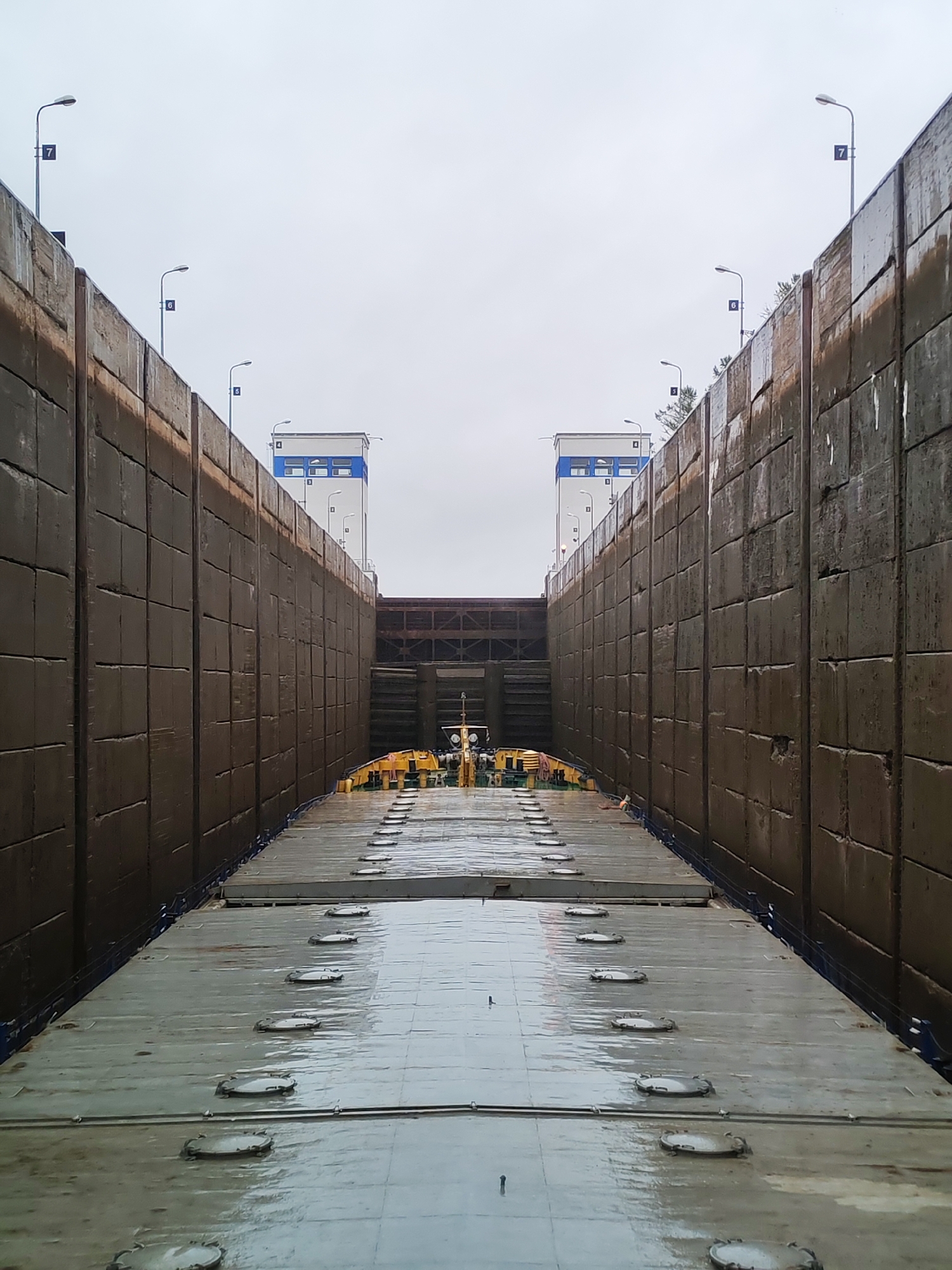 The image shows a view from the deck of a vessel inside a lock chamber. The lock walls are made of large, rectangular concrete blocks and rise high on either side. The deck of the vessel is flat, grey, and wet, with numerous circular hatches visible. In the distance, at the end of the lock, are two white structures with blue accents, and beyond them, a dark, cavernous opening that leads to the next section of the waterway. Several streetlights with numbered signs (7, 6, 5, and 4) are visible along the tops of the lock walls. The sky is overcast and a uniform grey. The scene appears to be in Devyatiny, Russia, likely at a canal lock system. No people are visible in the image.