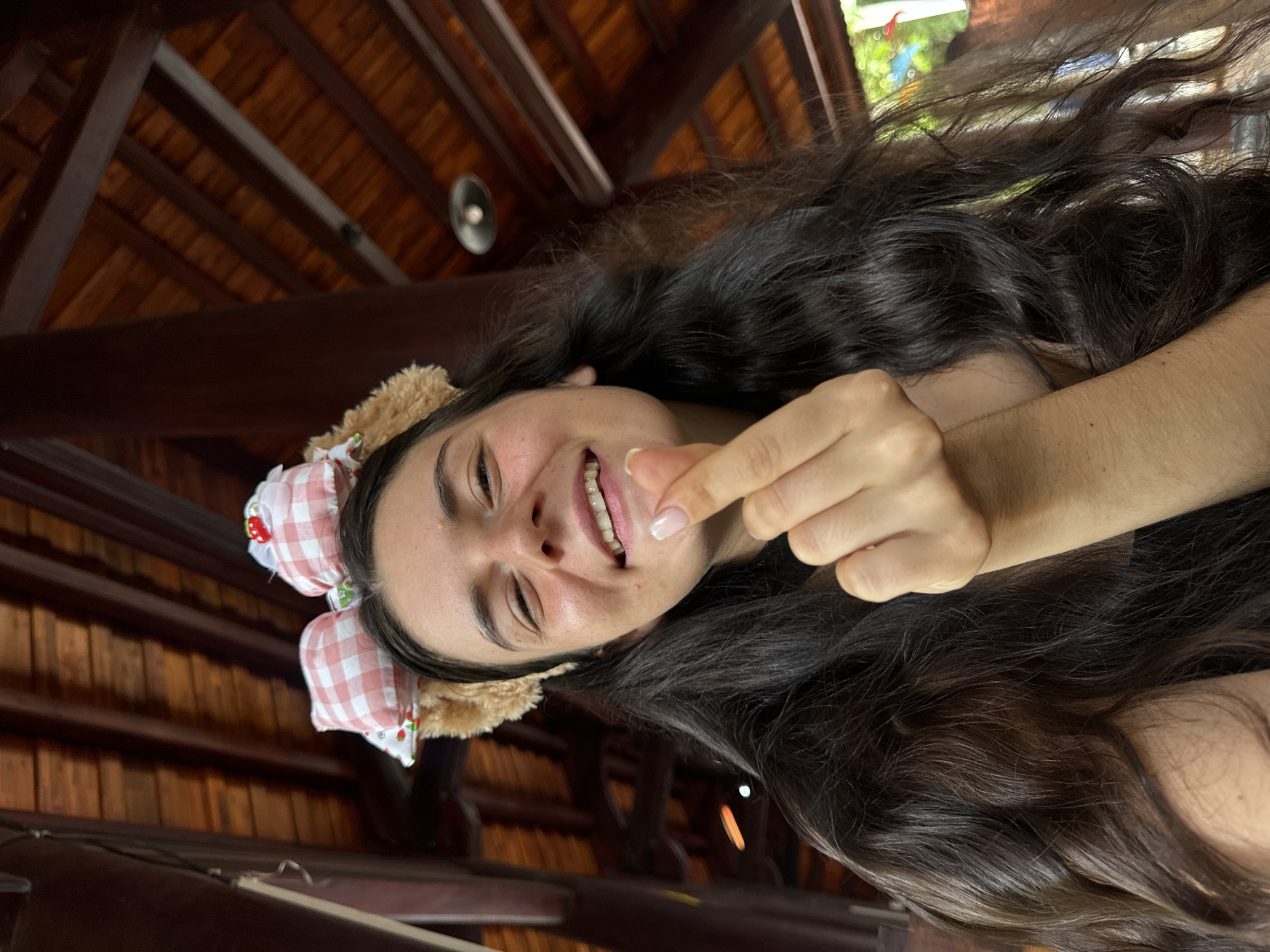 A young woman with dark, wavy hair and a pink and white gingham bow headband with a strawberry detail is smiling at the camera. She is wearing braces and making a heart shape with her fingers. The background appears to be an outdoor covered area with a wooden beamed ceiling. It is unclear if this location is in Mũi Né, Vietnam.