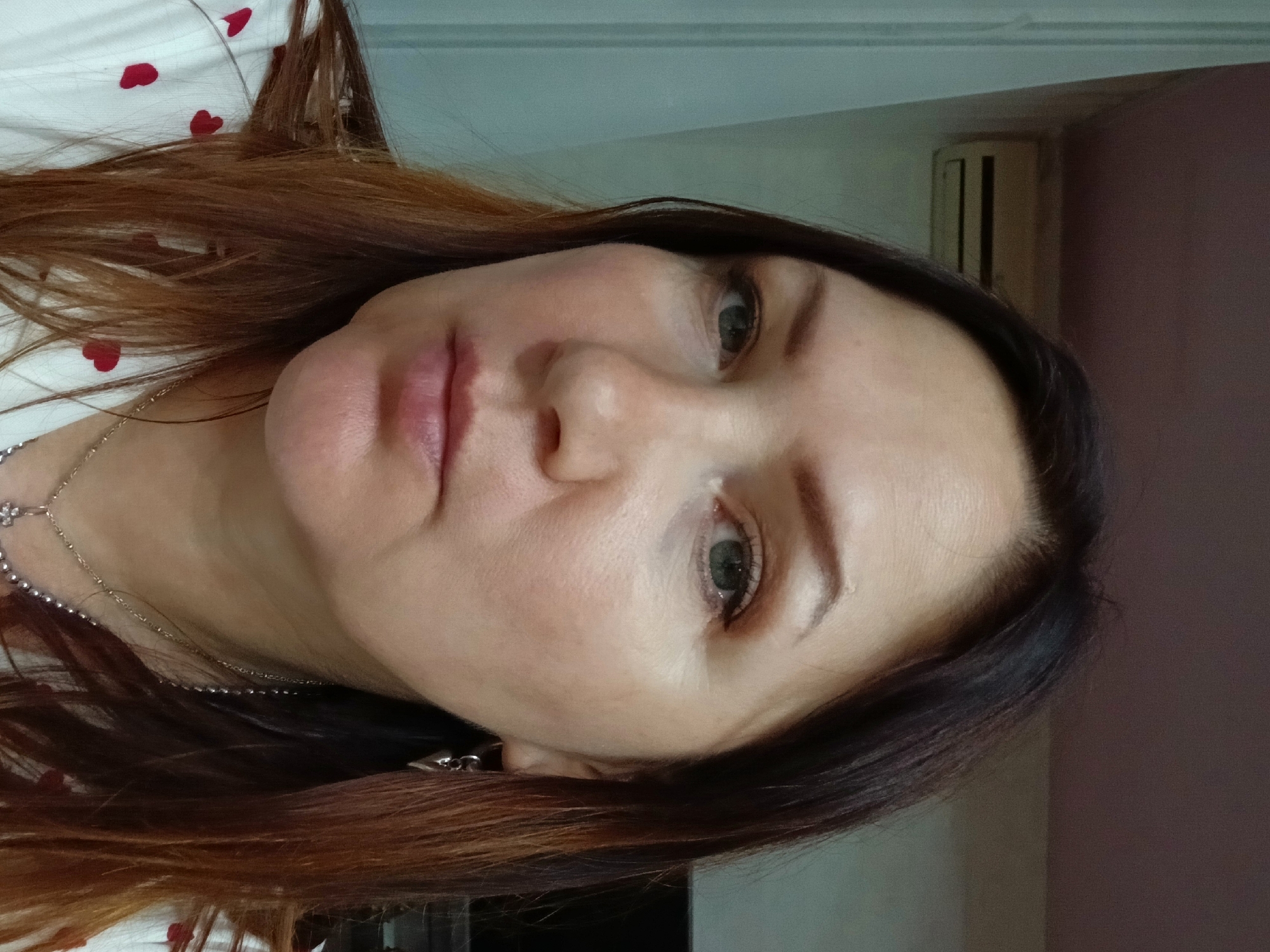 A woman with medium-length brown hair and fair skin is shown in a close-up selfie. She is wearing a white shirt with a red heart pattern, a silver necklace with a pendant, and a silver earring. Her expression is neutral. The background appears to be an indoor setting, possibly a room in Chaikovsky, Russia, with a wall, a curtain, and what looks like an air conditioning unit visible.