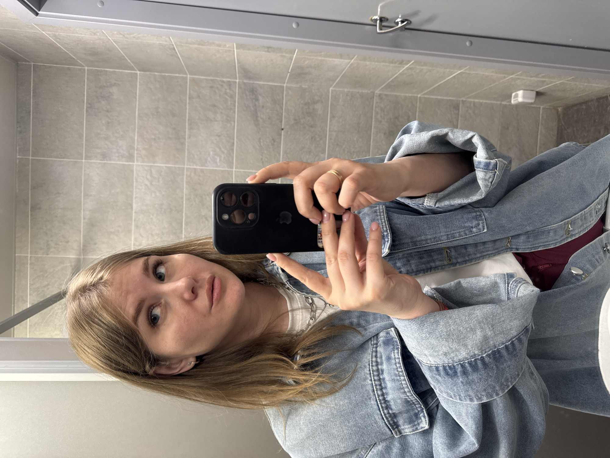 A young woman is taking a selfie in a bathroom. She is holding a black smartphone with multiple lenses and looking at the camera with a neutral expression. She has blonde hair and is wearing a denim jacket over a white top and a silver chain necklace. Her fingernails are painted light pink. A gold ring is on her right ring finger.
The bathroom has gray tiled walls and a mirror above a white countertop. Above the mirror are metal fixtures. A white rectangular object is visible on the wall near the upper right corner.
The location is Perm, Russia.