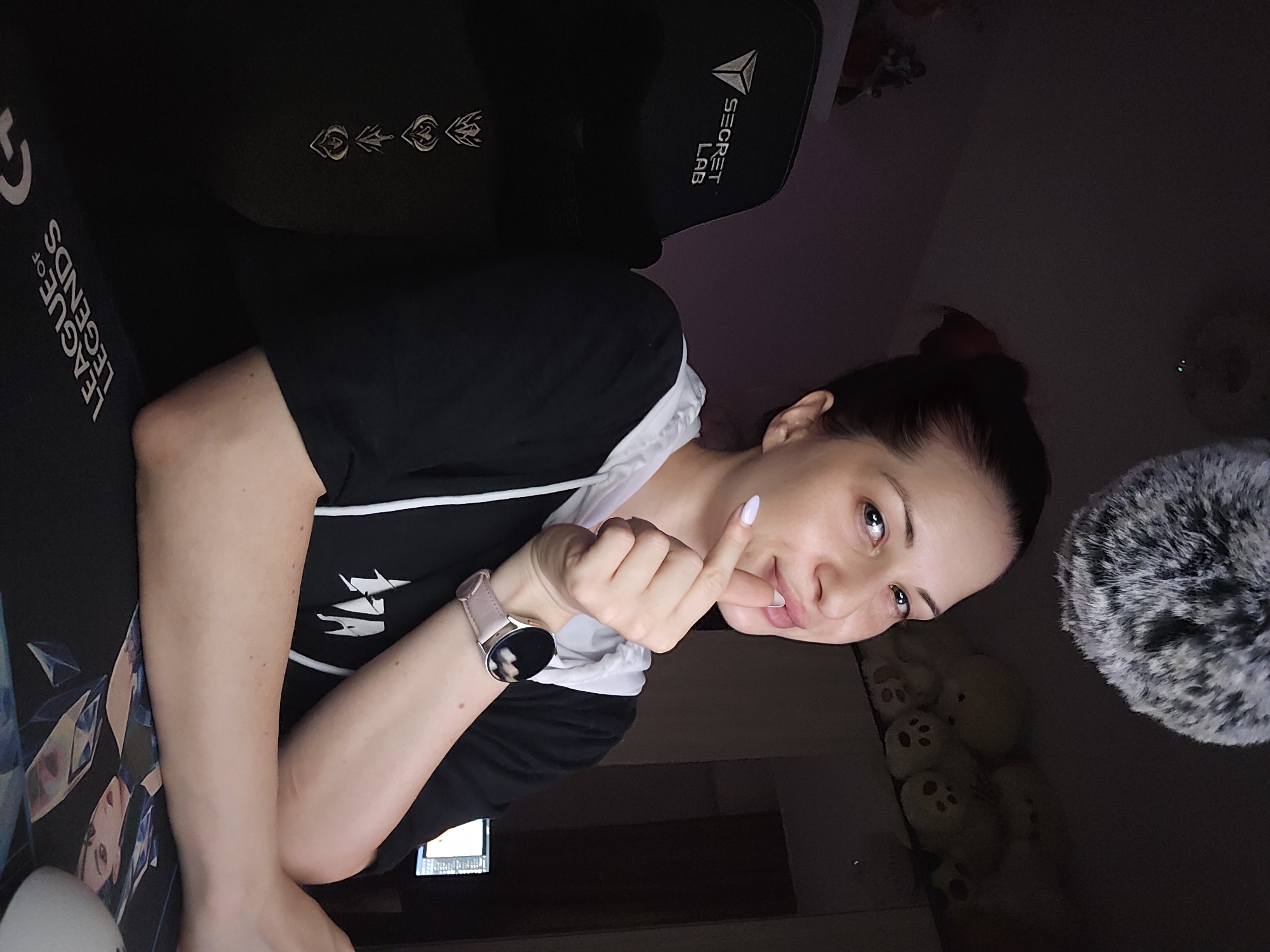 A young woman is pictured indoors, seated in what appears to be a gaming setup, likely at a desk. She is looking directly at the camera with a gentle smile, her right index finger playfully touching her lips. Her dark hair is pulled back, and she wears a black shirt or hoodie with white trim. A light-colored smartwatch with a circular face is visible on her left wrist, and her fingernails have a pale, possibly lavender, polish.
The setting is an indoor space, dimly lit. Behind her is a black gaming chair with the brand name "SECRET LAB" visible, along with embroidered symbols and partial text "LEGENDS" which likely refers to "League of Legends". To her left, a mousepad or desk mat is visible, prominently featuring "LEAGUE OF LEGENDS" text and anime-style character art. To her right, a fluffy, grey and white microphone windscreen is present. Below the microphone, several light-colored plush toys with embroidered faces are stacked. The overall environment suggests a gaming room or personal space. The woman's pose and direct gaze convey a friendly and possibly playful emotion. No specific external location cues are visible in the image itself, though the context indicates Burgas, Bulgaria.