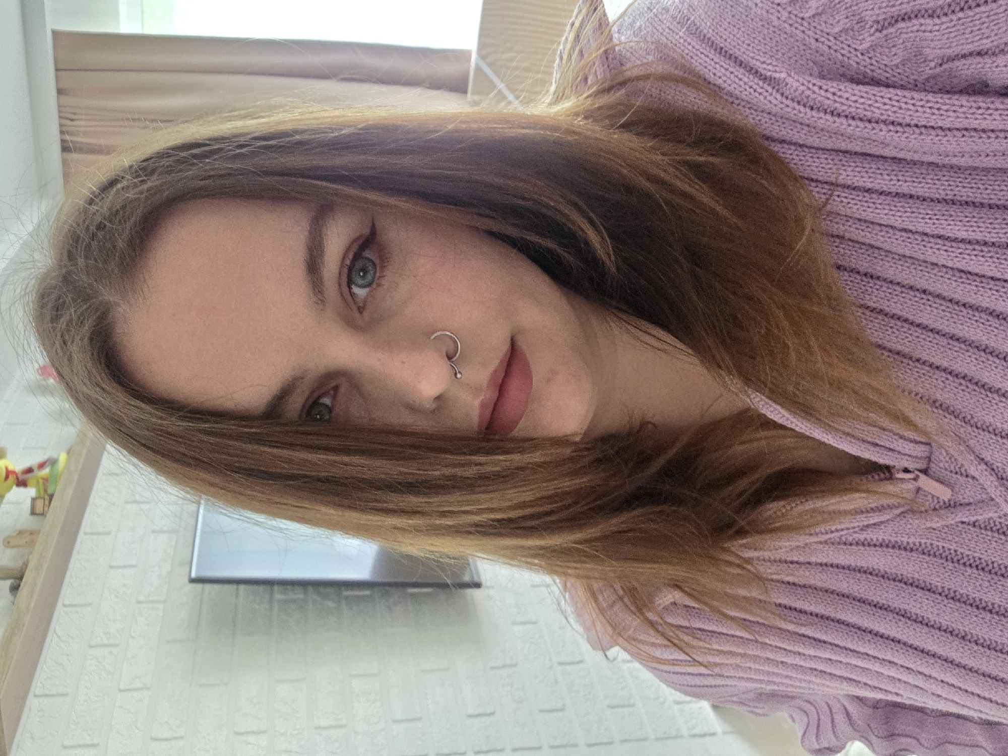 A young woman is angled toward the camera, wearing a lilac, ribbed sweater. She has light brown hair that partly covers her face, light-colored eyes, eyeliner, a nose ring, and pink lips. The background includes a window with closed blinds, a white textured wall, and a shelf with colorful toys. The location is in the city of Unknown, Unknown.