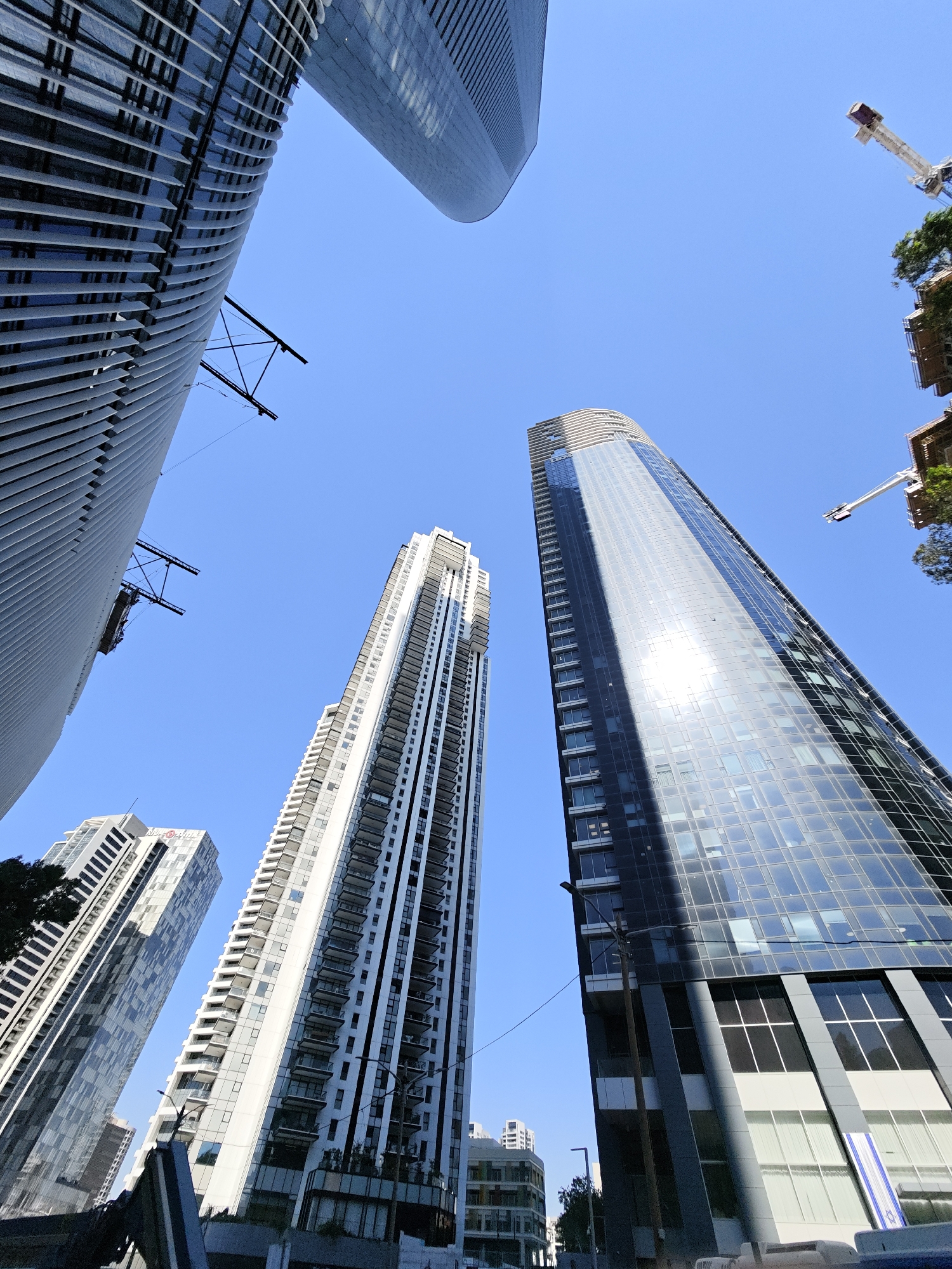 The photograph captures a low-angle view of several modern skyscrapers against a clear blue sky. The buildings are characterized by sleek, contemporary architectural designs, featuring extensive use of glass and metal facades.
In the foreground and left side of the image, a tall, curved building with a distinctive ribbed exterior is prominent. To its right, a slender, white skyscraper with numerous balconies and windows rises vertically. Further to the right, a dark, reflective skyscraper dominates the scene, its glass exterior mirroring the sky and surrounding structures. In the background, other buildings of varying heights and designs are visible, suggesting a dense urban environment.
The scene appears to be a construction or renovation site, indicated by the presence of a construction crane at the upper right portion of the image. The overall impression is one of urban development and modern architecture.
The location is identified as Givatayim, Israel, an urban area known for its mix of residential and commercial buildings, often featuring contemporary architectural styles. The clear sky suggests a sunny day, typical of the region's climate. No people are visible in the frame, focusing the attention solely on the architectural elements and the urban landscape.