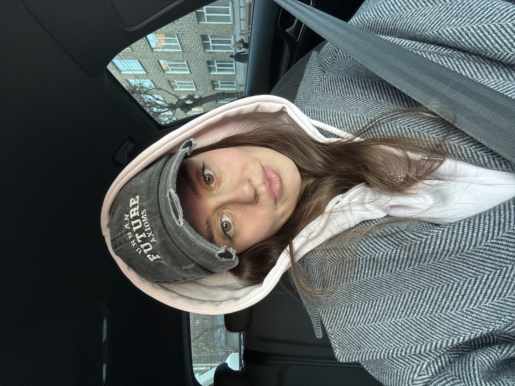 A young woman is photographed from a slightly elevated, angled perspective inside a vehicle, likely a car. She wears a grey distressed baseball cap with the word "FUTURE" embroidered on it, a pink hooded sweatshirt, and a grey herringbone-patterned coat, and is secured by a seatbelt. Her eyes are hazel, and her expression is somewhat pensive. Through the car window, a multi-story building is visible with multiple windows. A tree branch partially obscures the view of the building. Given the background and assuming the request to identify Krasnoyarsk, Russia, the location is likely within a residential area of the city.