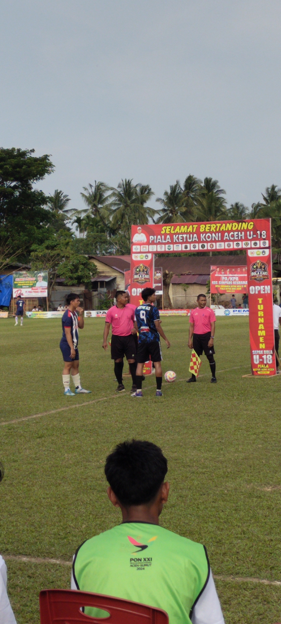 This image captures a soccer match in Keudesimpangempat, Indonesia. In the foreground, a young person wearing a bright green vest with the "PON XXI ACEH-SUMUT 2024" logo is seated on a red chair, their back to the camera. The middle ground shows a soccer field with a white boundary line. Three individuals are standing on the field: two men in pink shirts and black shorts, likely referees or officials, and a young man in a blue and white striped soccer jersey with the number 4 on his back. Another young man in a black and blue soccer jersey with the number 9 is also visible. A colorful soccer ball rests on the grass. Behind them, an archway with banners announces "SELAMAT BERTANDING PIALA KETUA KONI ACEH U-18" (Congratulations on competing, Koni Aceh U-18 Cup). The background features palm trees and simple buildings, indicating a rural or semi-urban setting. The sky is overcast.