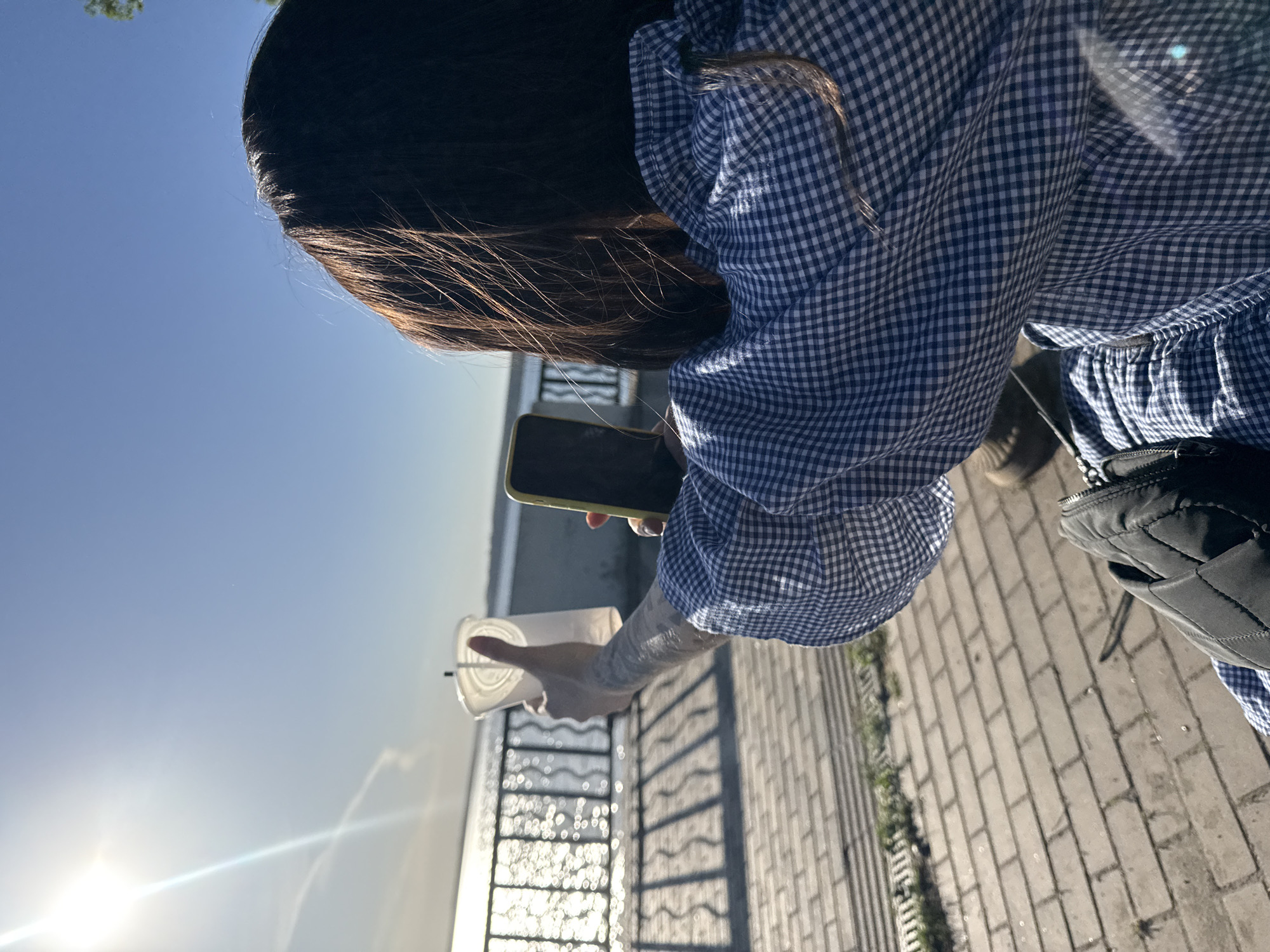 A person with dark hair is standing on a paved walkway next to a metal railing, holding a clear plastic cup in one hand and a smartphone in the other. The person is wearing a blue and white checkered dress. A black bag hangs at their side. The background shows a body of water under a clear blue sky with the sun shining brightly. The setting appears to be outdoors on a sunny day. It is not possible to confirm that the location is in the city of Votkinsk, Russia.
