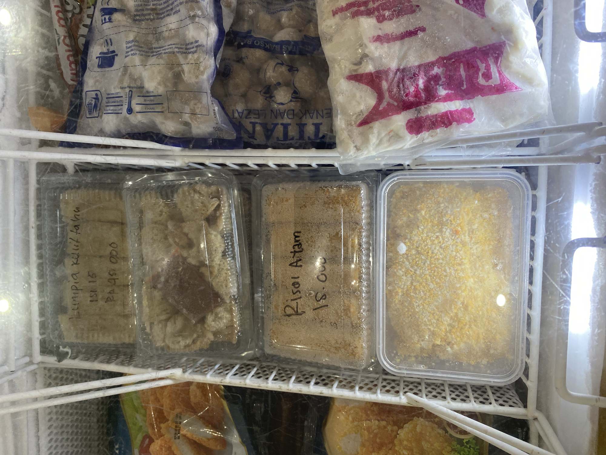 The image shows a freezer compartment filled with frozen food items. In the foreground, a white plastic basket contains several containers of frozen food. The top container has a handwritten label that reads "Lumpia Kulita Isi 15 Rp 45.000". The second container from the top appears to hold a dark, frozen substance. The third container from the top has a label that reads "Risol Ayam 15.000". The bottom container in the basket is filled with what appears to be breaded food items. To the right of the basket, there are several sealed plastic bags containing frozen food, one of which has the brand name "TITAN" visible. To the left of the basket, another plastic bag contains breaded food items. The freezer compartment itself is white and appears to be in a commercial setting, possibly a store or restaurant in Payatieng, Indonesia. There are no people visible in the image.