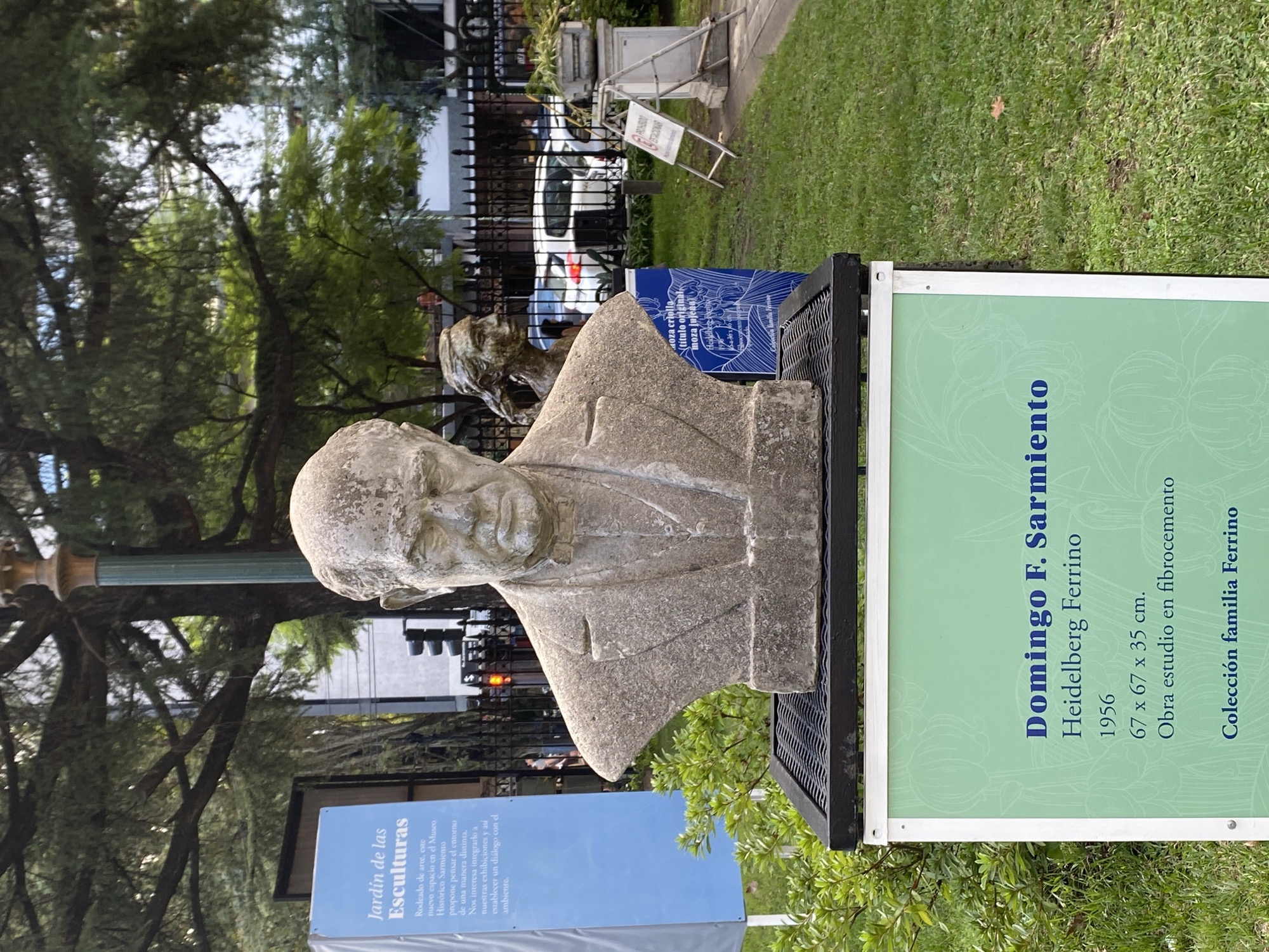 The image shows a bust of Domingo F. Sarmiento in a park setting. A green and white sign next to the bust identifies it as "Domingo F. Sarmiento" by Heidelberg Ferrino, created in 1956. The dimensions are listed as 67 x 67 x 35 cm, and it is described as a study piece in fiber cement, from the Ferrino family collection. Another sign indicates that this is the "Jardín de las Esculturas" (Sculpture Garden) and explains the purpose of the space in relation to the Museo Histórico Sarmiento. In the background, there are trees, a black iron fence, a parked car, and a sign. Based on the sign referring to the Museo Histórico Sarmiento, the location is likely in Buenos Aires, Argentina.