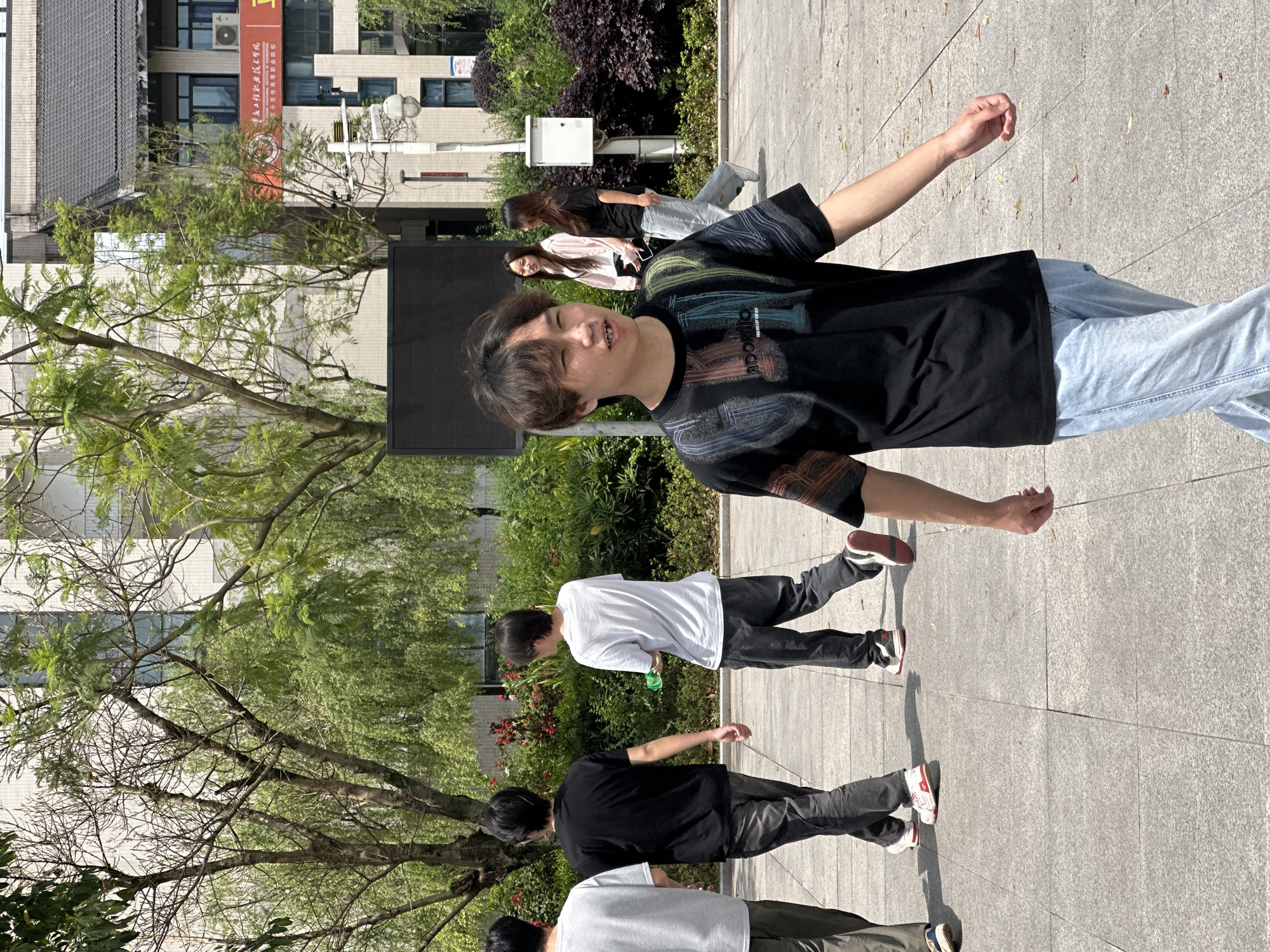 The image captures a moment on a paved outdoor space in Jiangjin, China. A young man in a black t-shirt and light blue jeans stands prominently in the foreground, with his arms extended. Behind him, a group of other individuals are visible, walking in the same direction. These include people wearing both light and dark shirts. The surrounding environment features trees, bushes, and a building.
