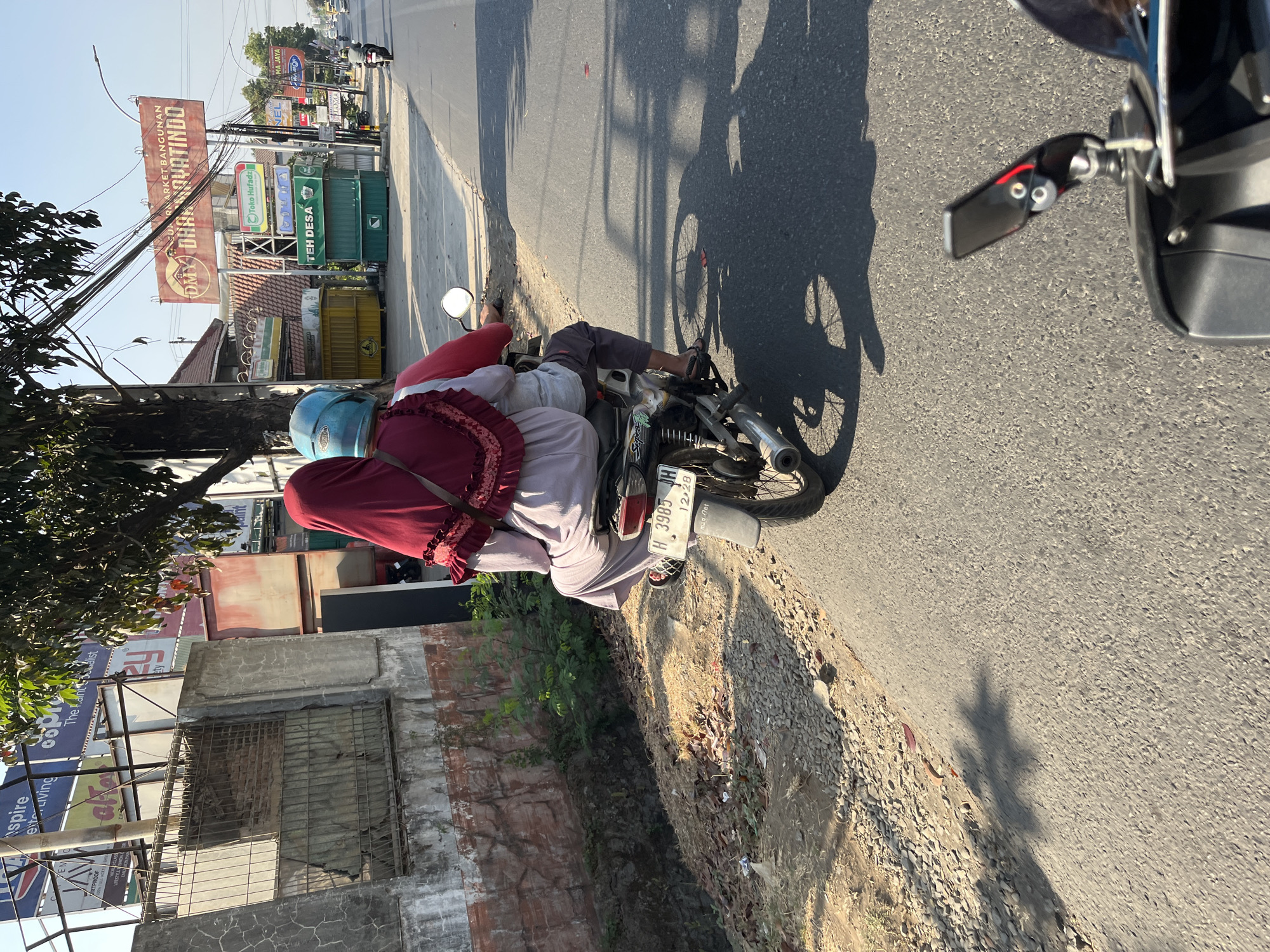 The image shows two people riding on a motorcycle on a street in Kalinyamatan, Indonesia. The person in the back is wearing a maroon hijab with ruffled trim and a light purple top, and the person in the front is wearing a blue helmet. The motorcycle has a license plate that reads "H 3985 JH". The environment is a street with buildings and billboards in the background. The sun is shining, casting shadows on the road.