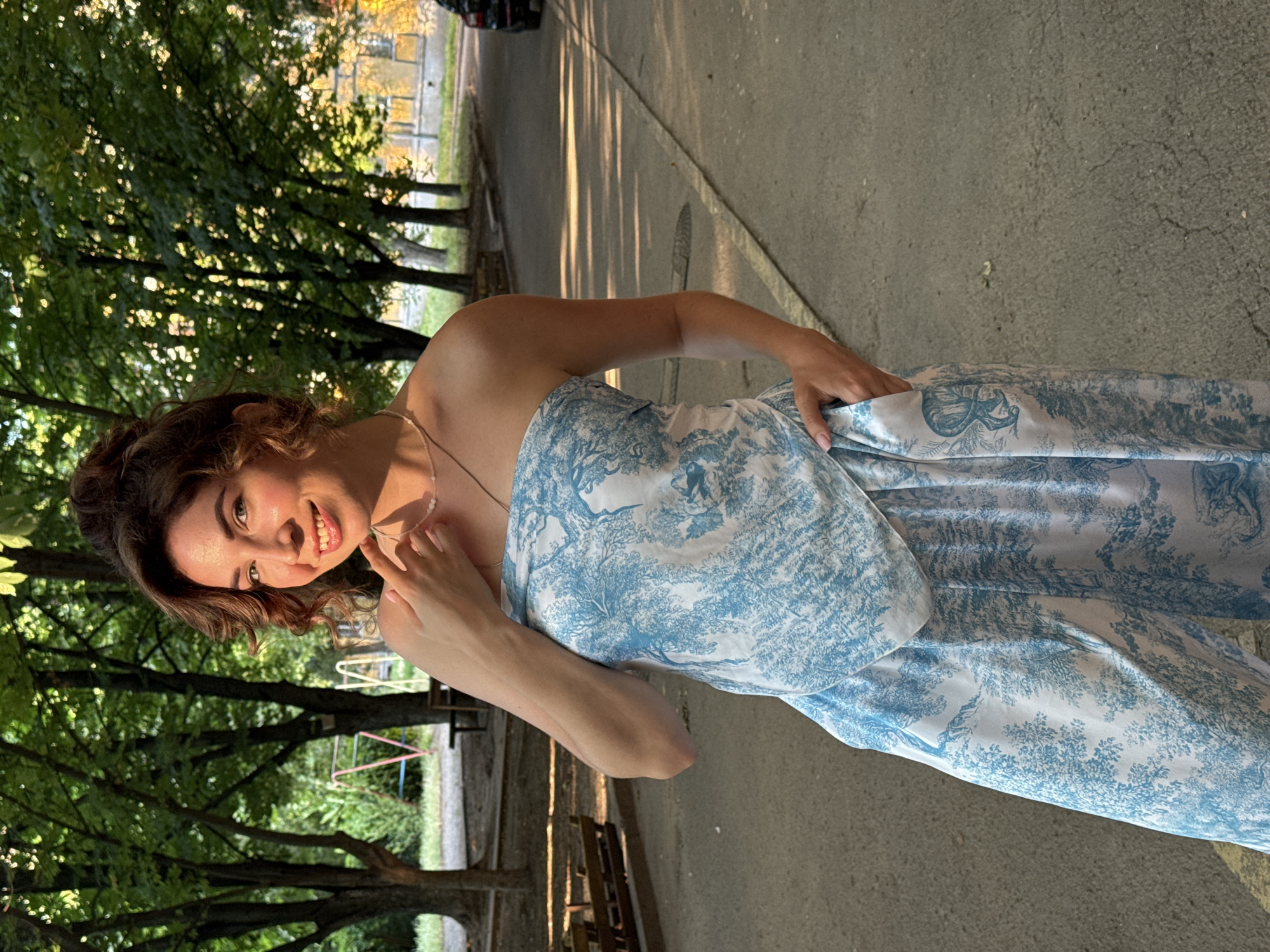 A young woman with curly brown hair and a bright smile poses for a photo outdoors. She is wearing a strapless dress with a blue and white toile pattern. Her left hand is tucked into her dress, and her right hand is near her face, with her fingers touching her chin. She is looking directly at the camera.
The background shows a paved walkway or road lined with trees. Sunlight filters through the leaves, casting dappled shadows on the ground. In the distance, playground equipment and buildings can be seen. The scene appears to be set in a park or residential area.
The lighting suggests it is daytime, likely late afternoon or early evening, given the warm, angled sunlight. The woman's expression is joyful and engaging, conveying a sense of happiness and confidence. The overall mood of the image is cheerful and relaxed. No visible text is present in the image.