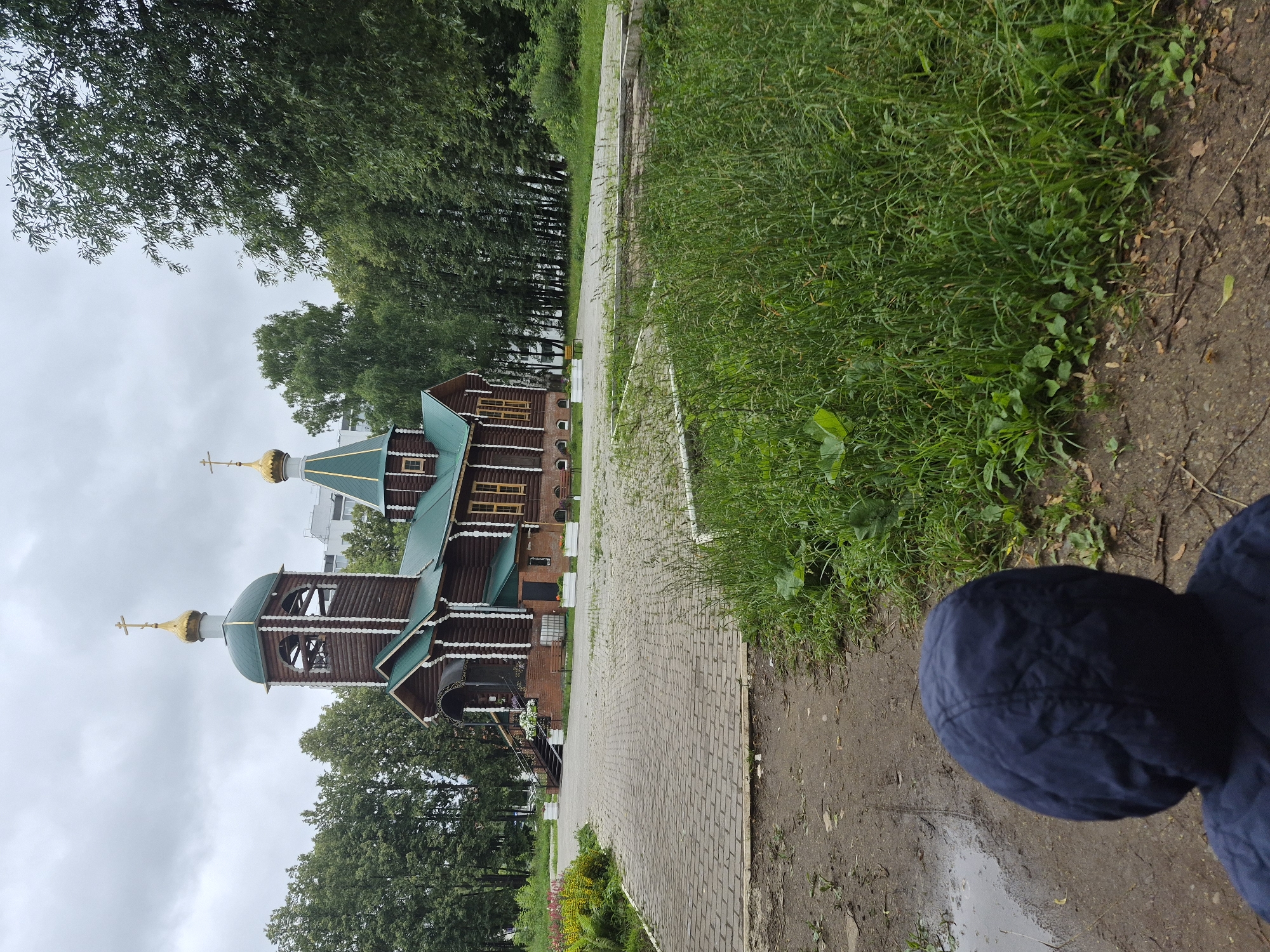 A wooden Orthodox church with green roofs and golden domes stands amidst lush greenery under an overcast sky. The church features a bell tower to the left and an entrance with a staircase. A paved walkway leads towards the church, bordered by grass and some flowering plants. In the foreground, the back of a person wearing a dark blue hooded jacket is visible, facing away from the camera and towards the church. The ground in the foreground is a dirt path with some puddles. The location is the Church of the Intercession of the Most Holy Theotokos in Chaikovsky, Russia.