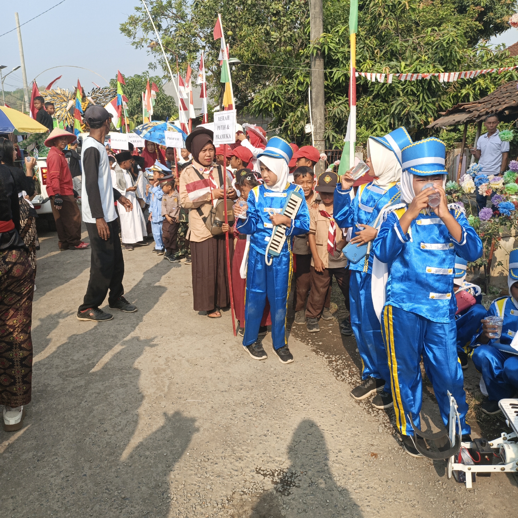 In Tanahbaya, Indonesia, a group of children in uniform are participating in a parade. The children are dressed in bright blue uniforms with yellow stripes, some wearing blue hats with yellow trim. Many of them are holding musical instruments, such as a keyboard. Some of the children are drinking from clear plastic cups. Several adults are also present, observing the parade. The scene takes place on a paved road, with trees and buildings visible in the background. Flags and banners, some with red and white Indonesian colors, are displayed along the parade route. The lighting suggests it is a sunny day.